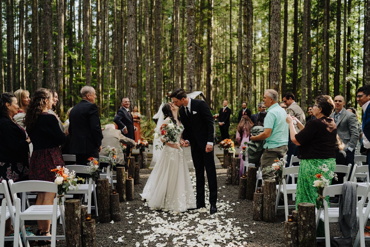 Forest wedding ceremony with guests seated on white chairs among tall pine trees.