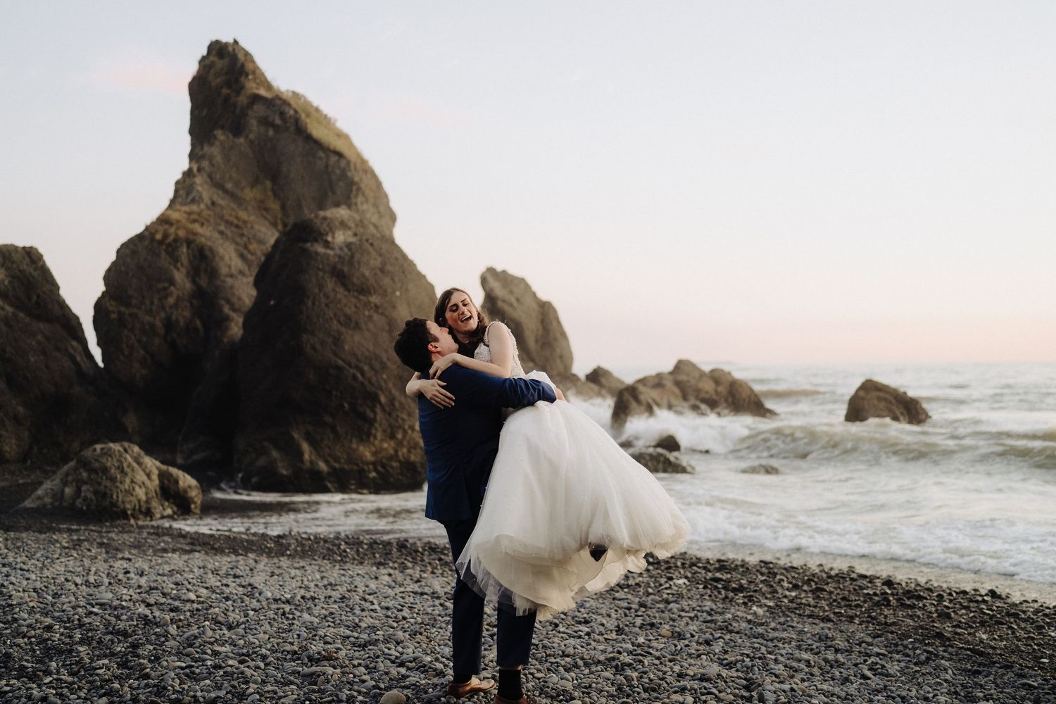 Wedding couple poses on rocky beach with dramatic sea stacks and ocean waves at dusk.