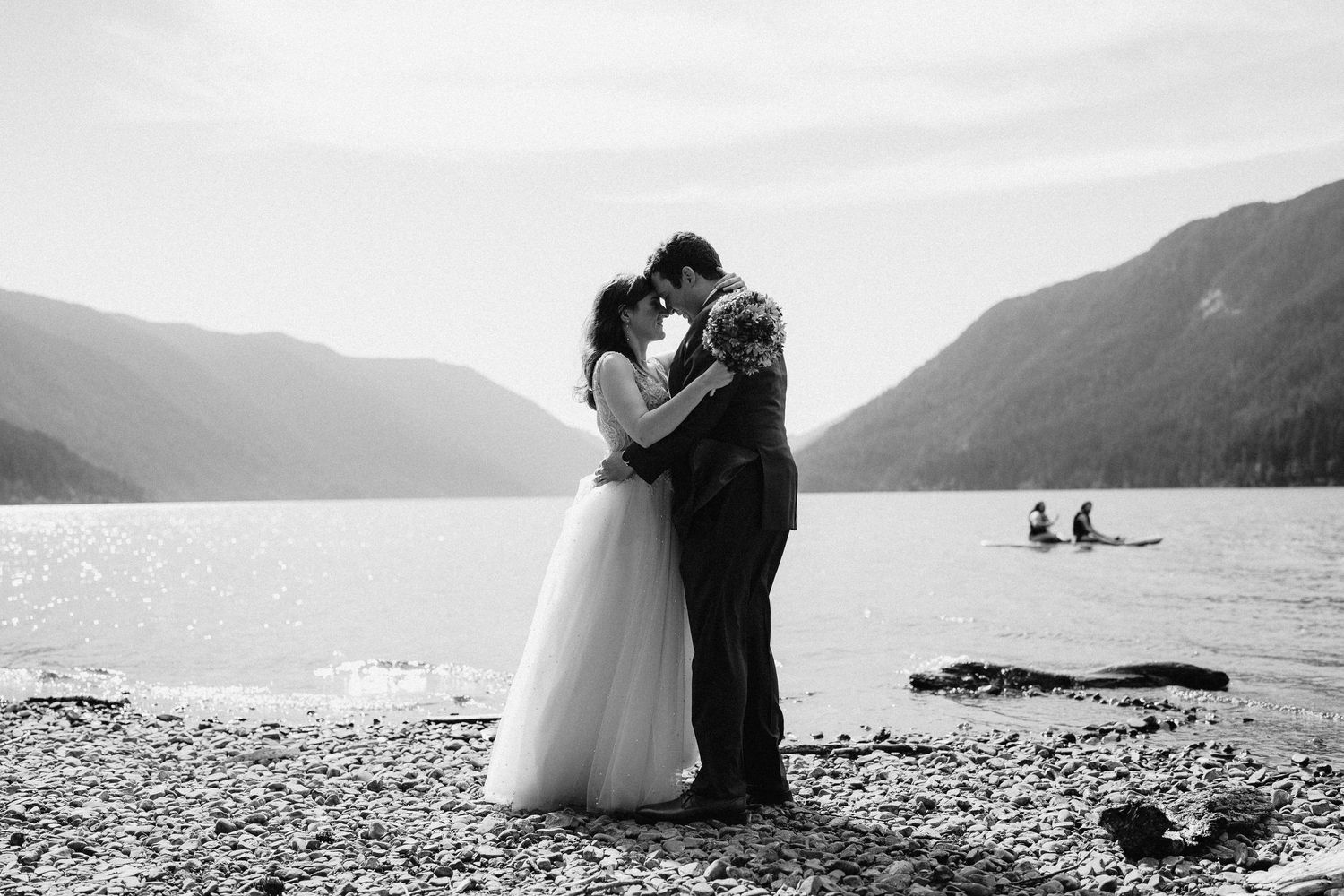 A black and white wedding photo captures an embrace by the lake with mountains in the distance.