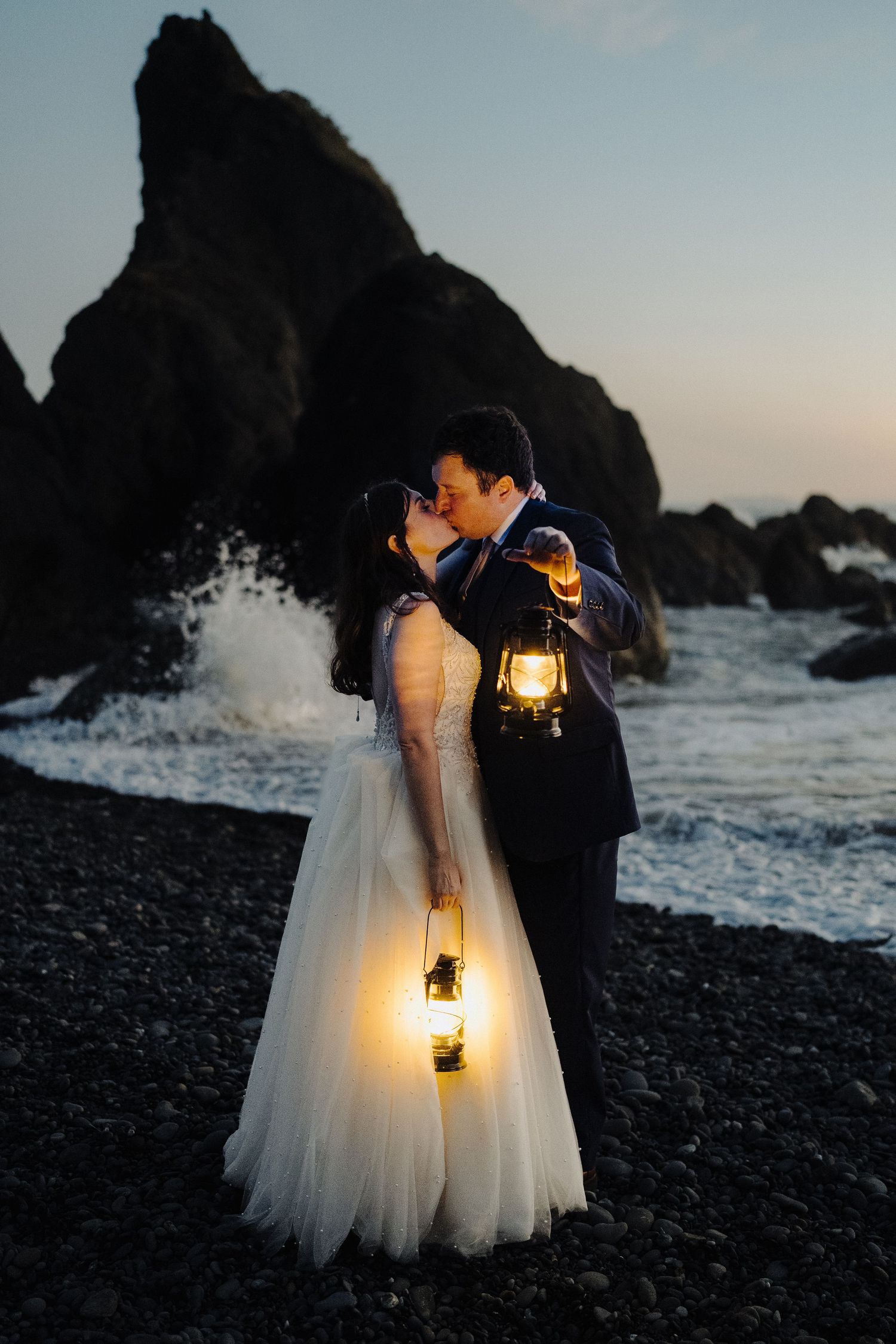 A couple shares a romantic moment with lanterns on a dark beach beside towering sea stacks and crashing waves.