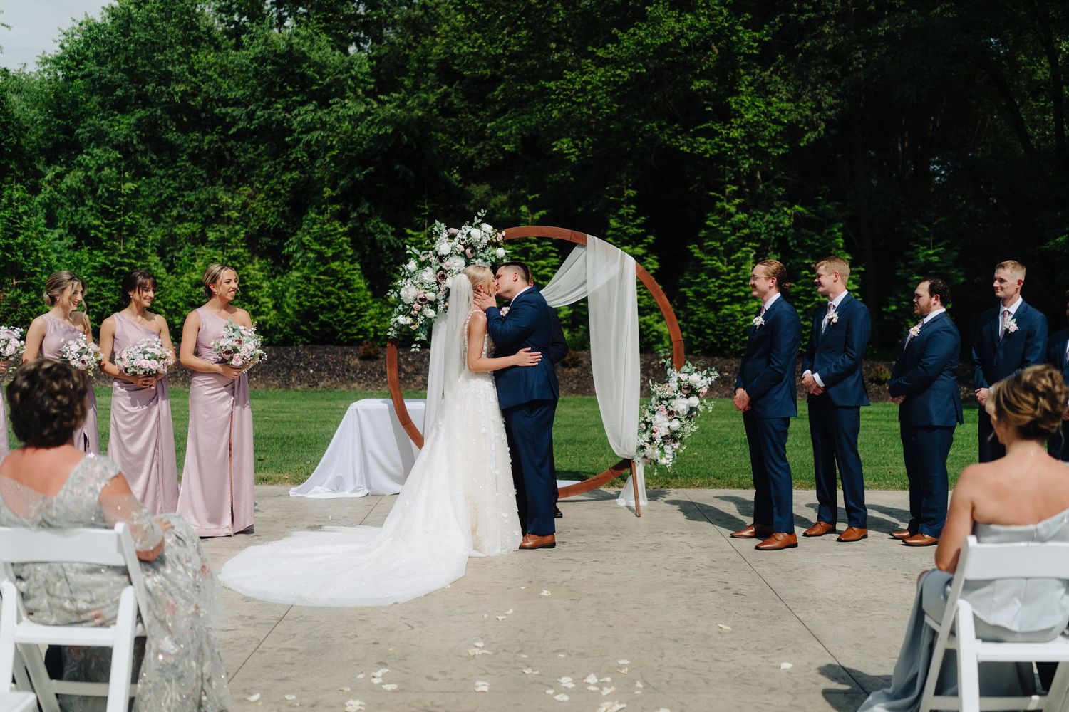 Outdoor wedding ceremony setup with white chairs and floral arch under green trees.