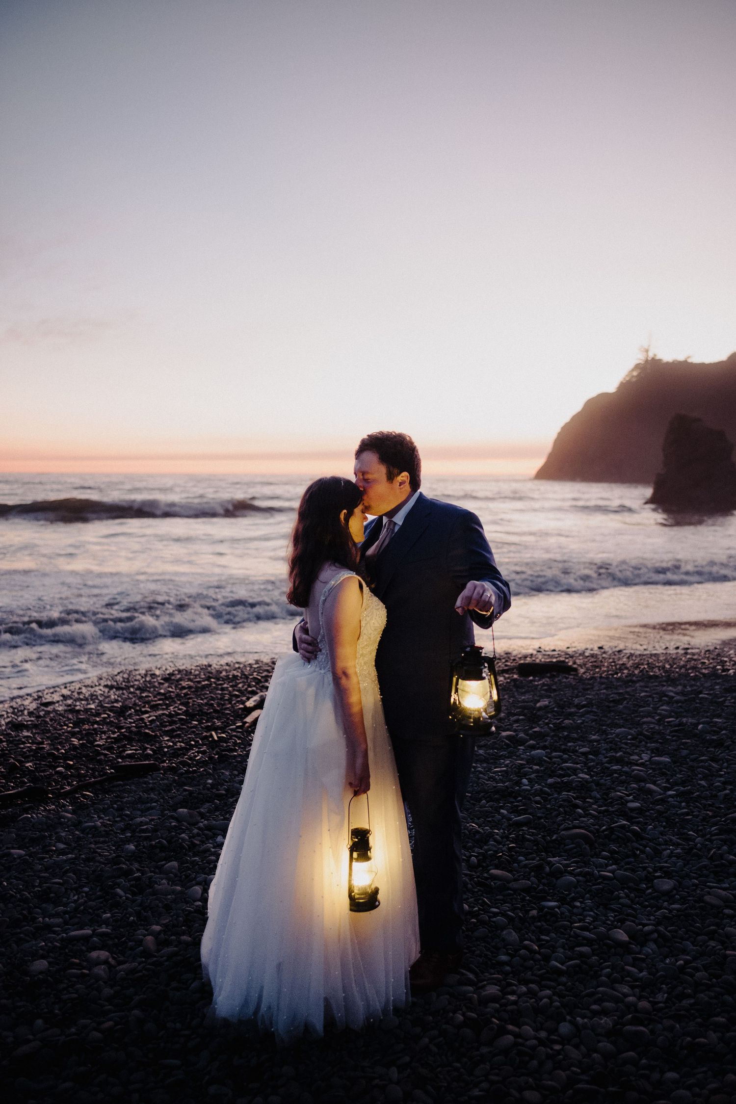 A romantic beach wedding portrait at sunset with lanterns and crashing waves in the background.