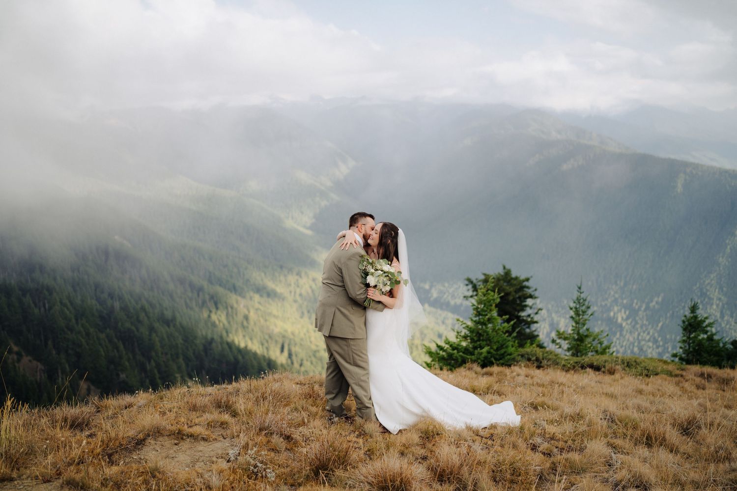 A couple shares an intimate moment on a mountaintop overlooking a misty mountain range and lush green valleys.