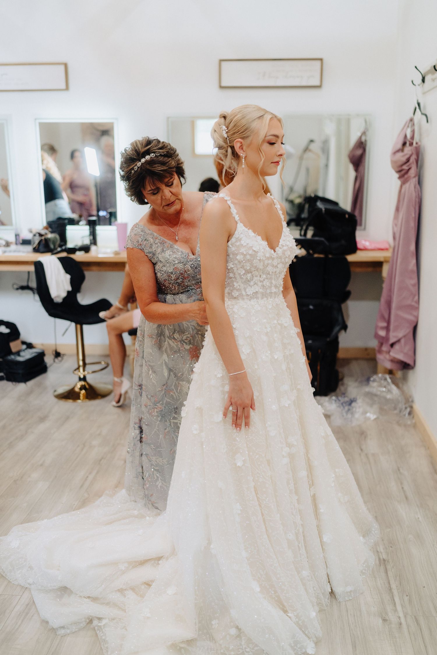 A bride in a white lace gown shares an emotional moment with someone wearing a silver sequined dress in a bridal boutique.