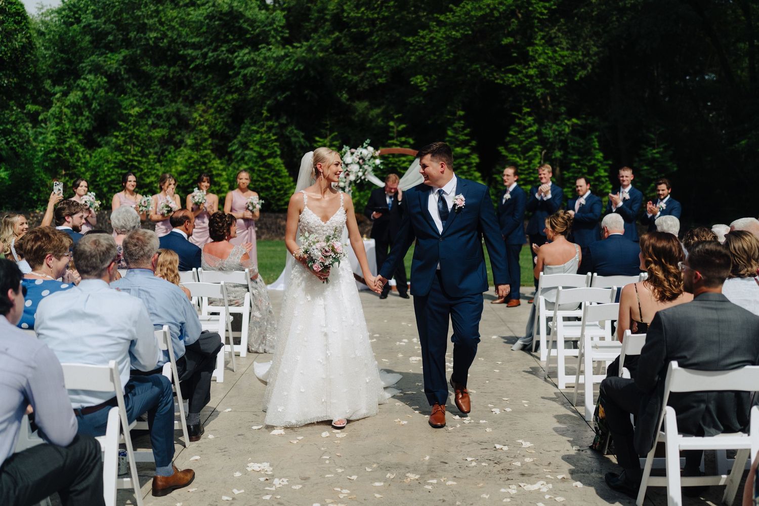 Wide angle shot of wedding ceremony with guests seated in white chairs surrounded by trees.