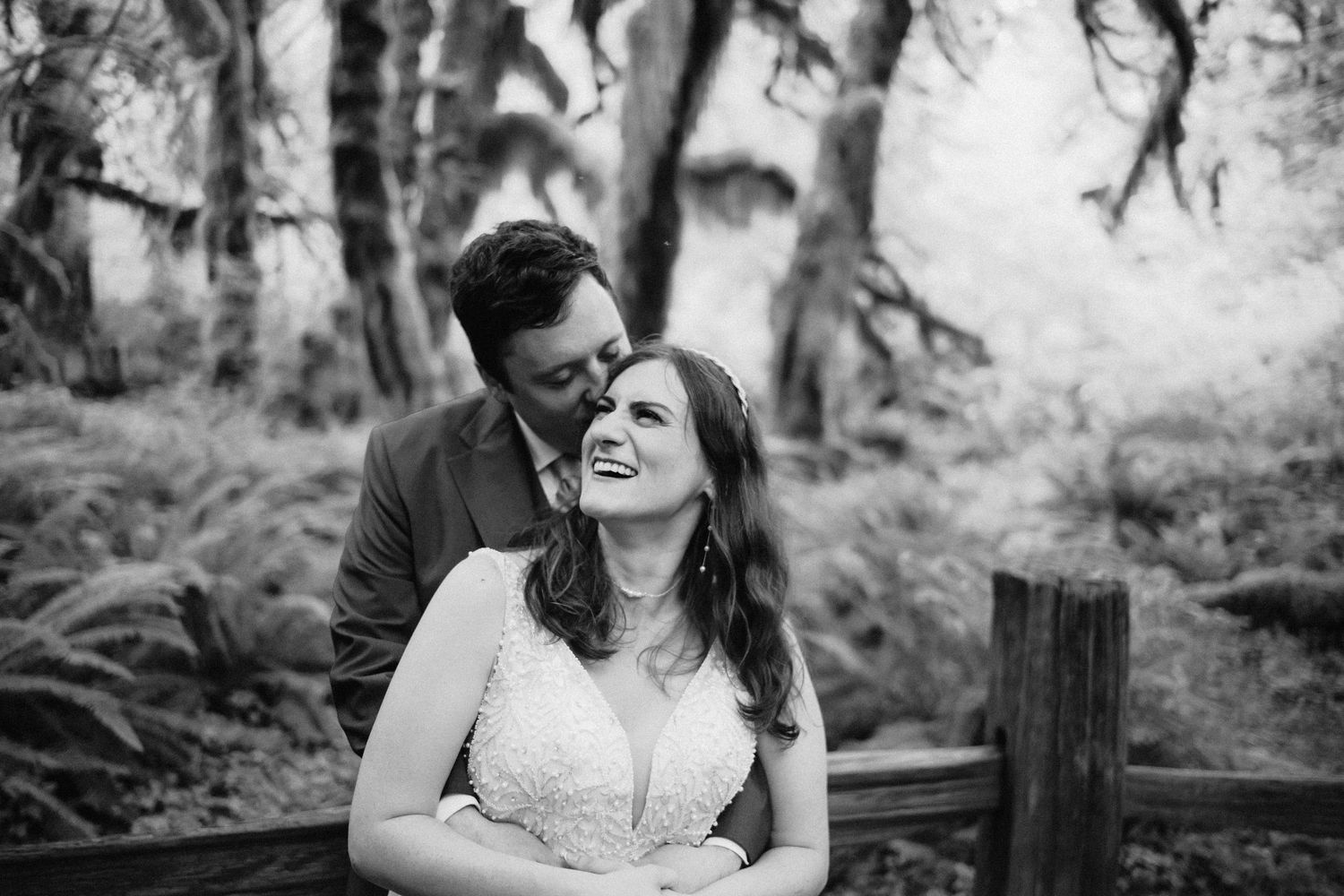 A black and white portrait of a couple sharing a joyful moment on a forest boardwalk.