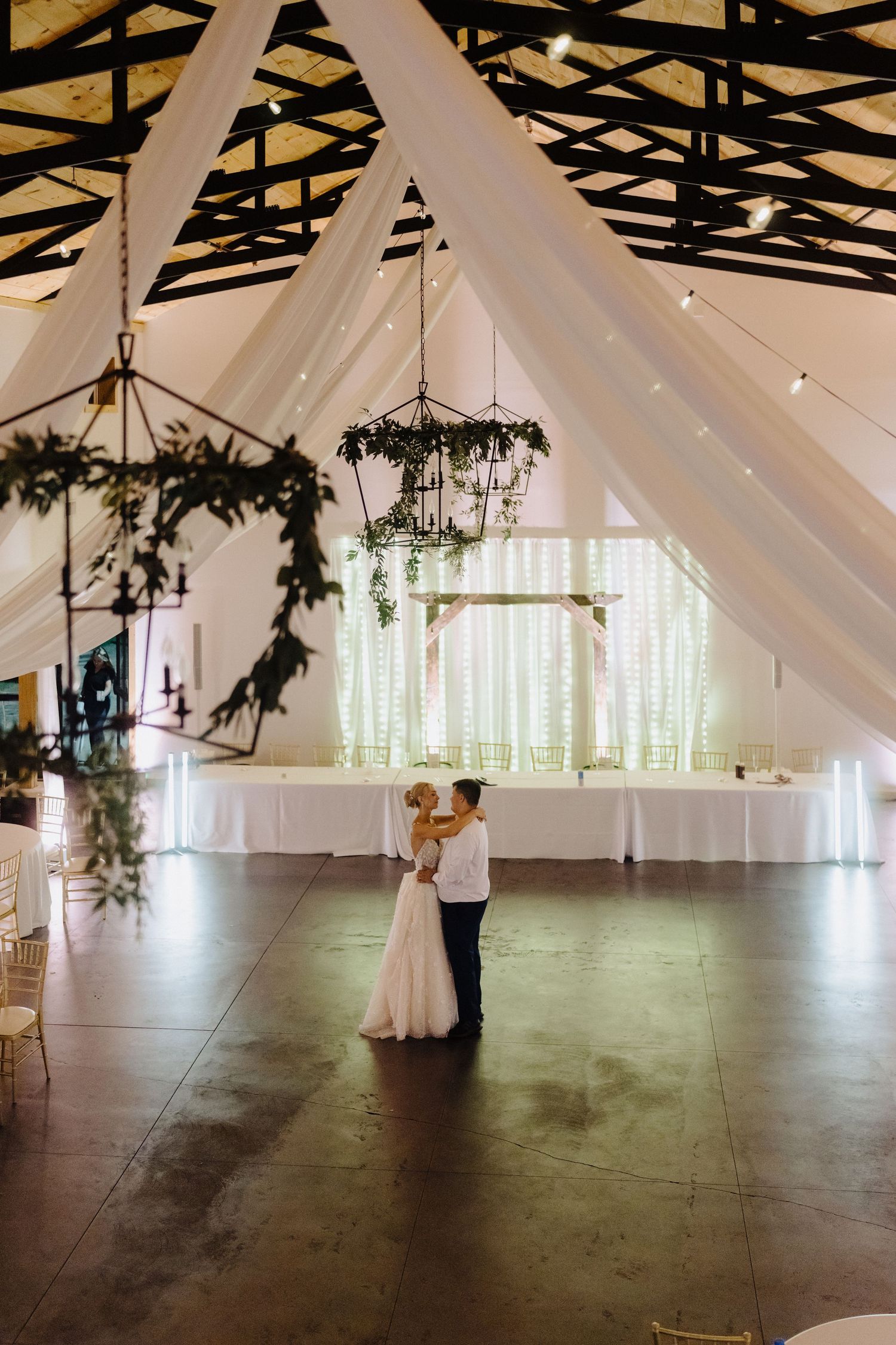 An elegant white wedding venue with draped fabric and greenery hanging from exposed wooden beams.