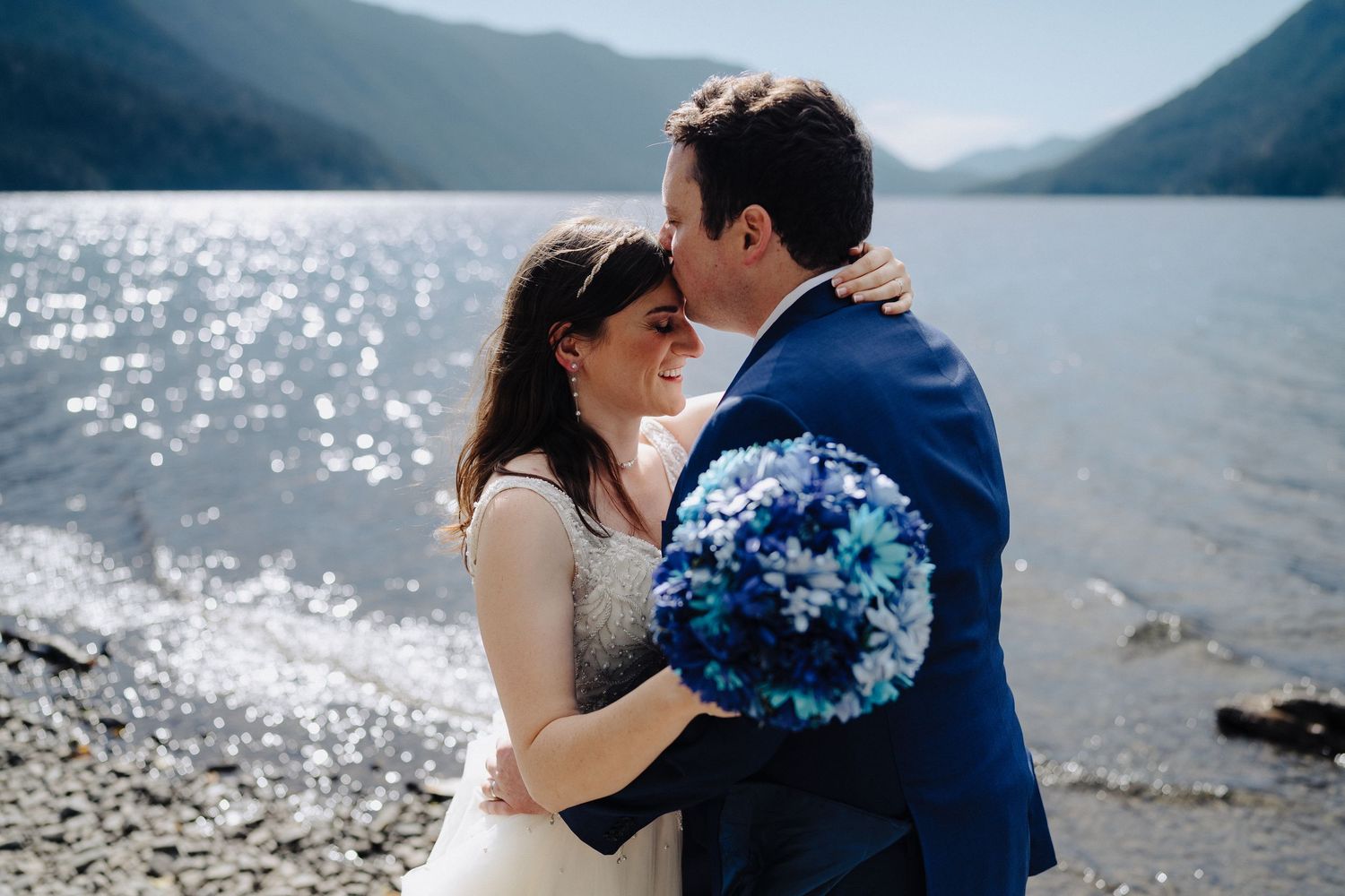A romantic moment captured on a sunny lakeside beach with sparkling water and mountains in the distance.