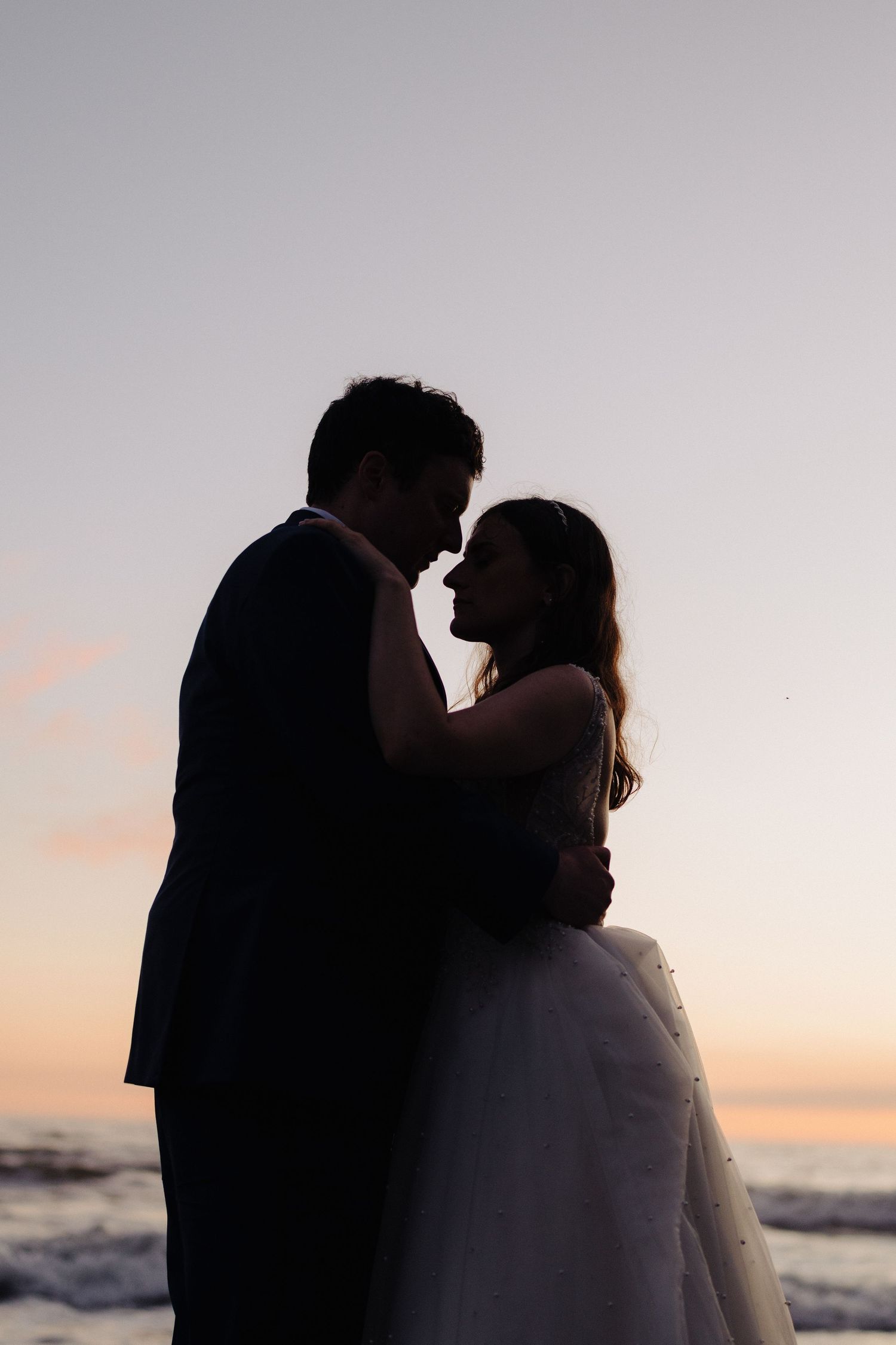 Silhouette of a bride and groom standing close together against a sunset sky by the ocean.