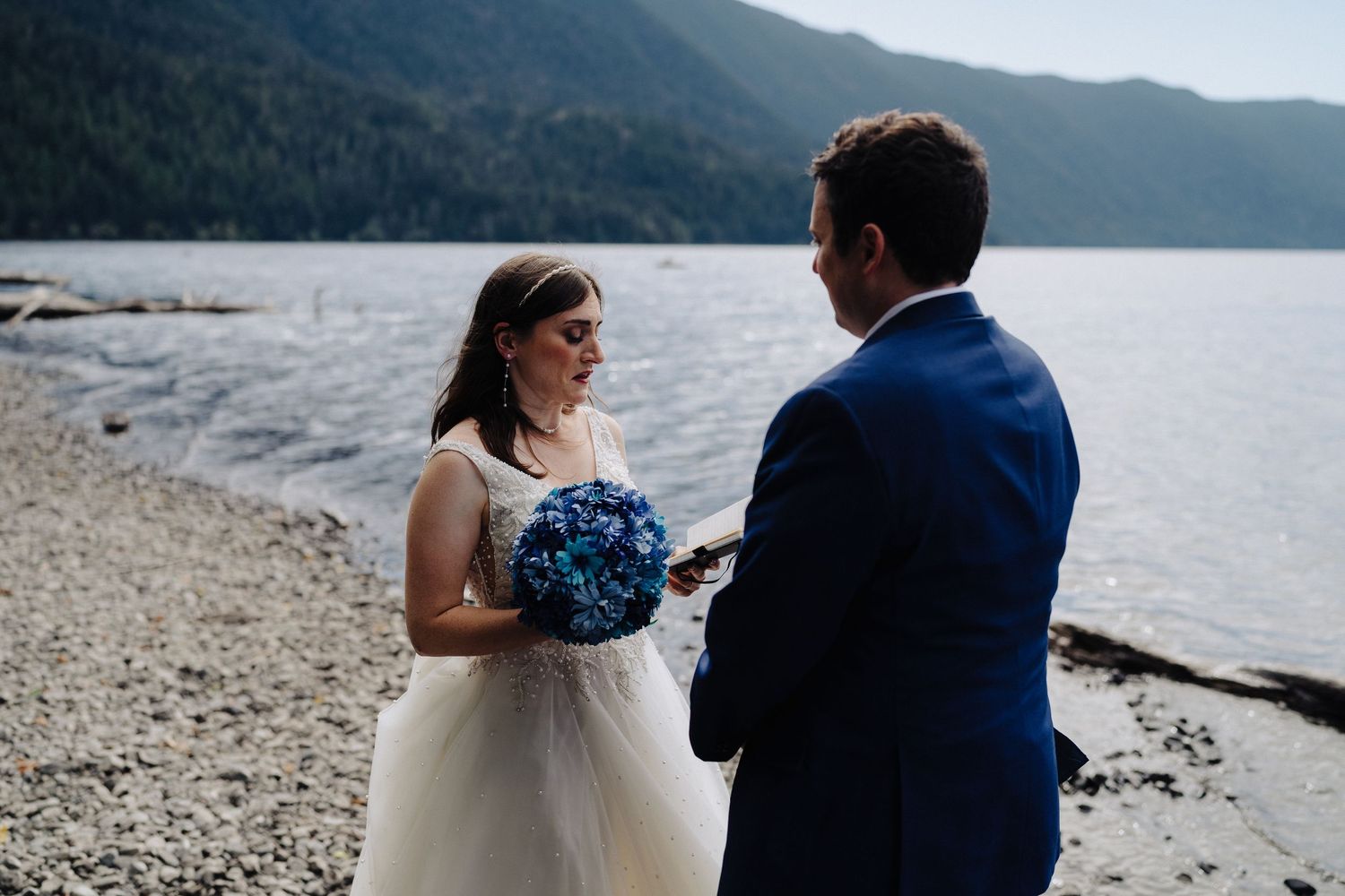 Wedding couple exchanges vows on a pebble beach with mountain lake scenery.