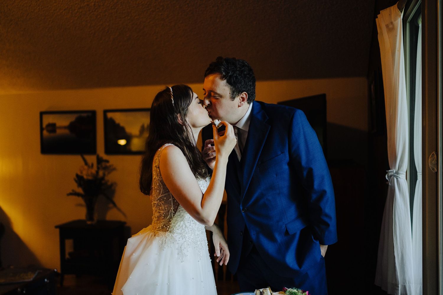 A couple in wedding attire share an intimate moment in a dimly lit room next to a window.