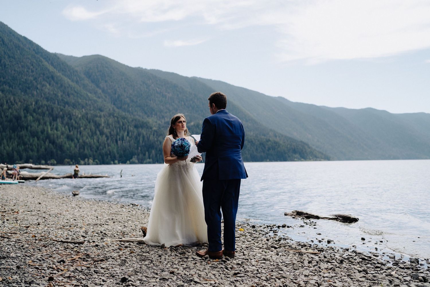 A wedding couple stands together on a rocky lakeshore surrounded by mountainous landscape.