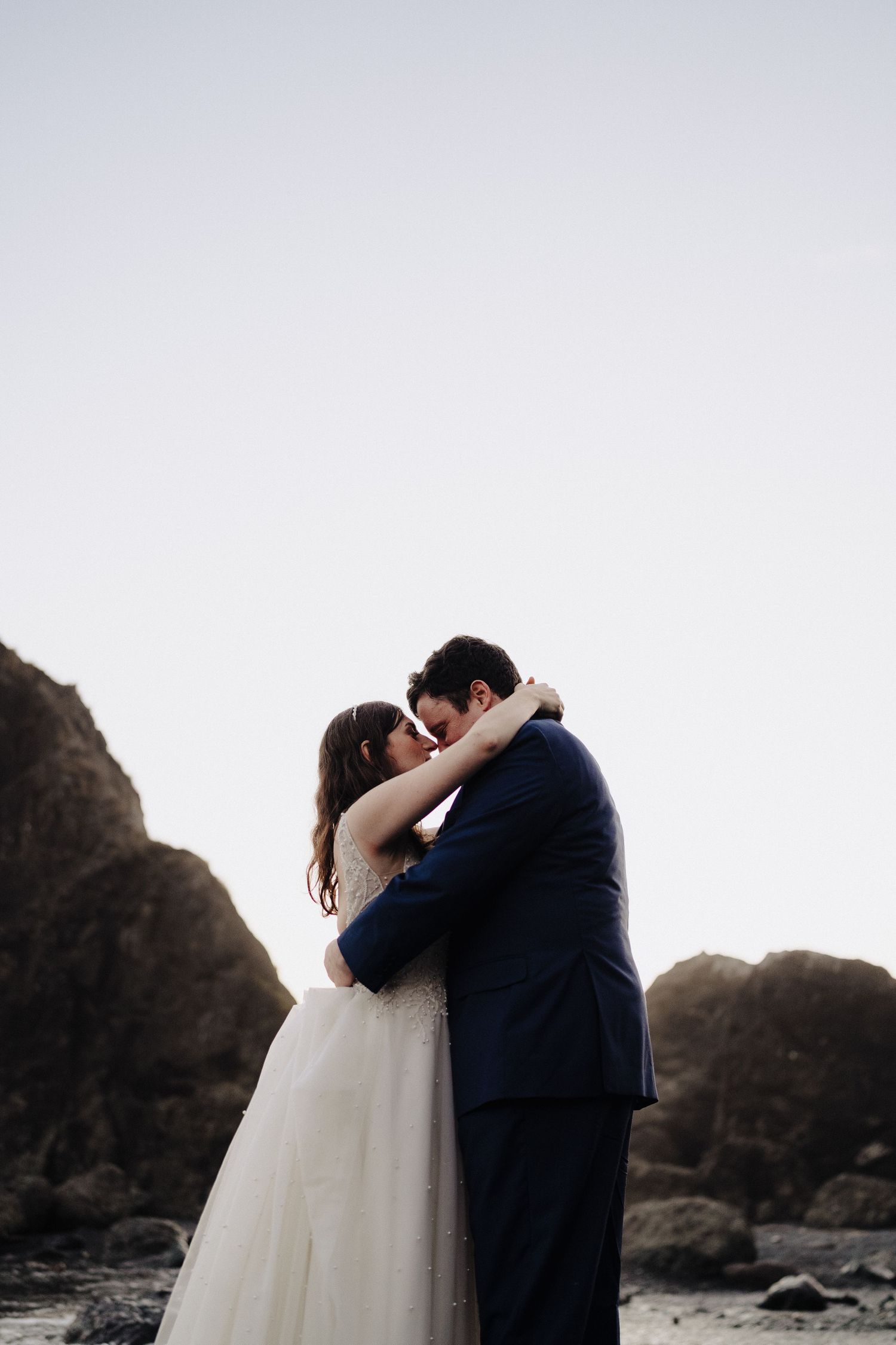 A romantic beach embrace between newlyweds against dramatic rocky cliffs at sunset.