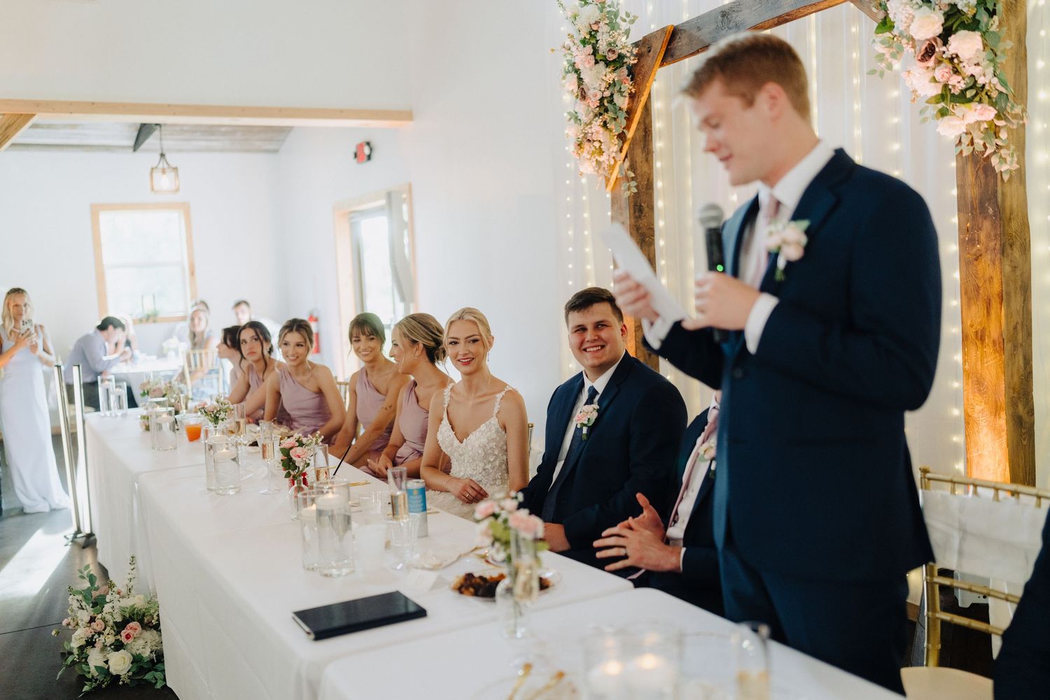 Wedding reception venue decorated with floral arrangements and white draping in a rustic indoor space.