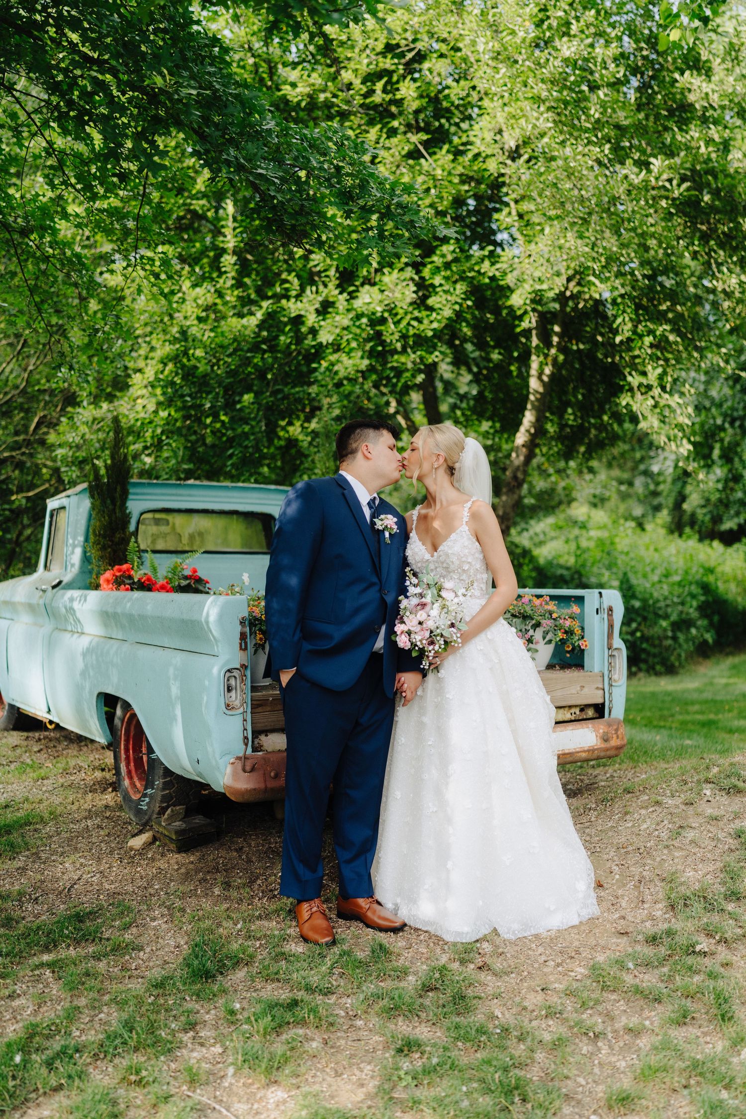 A newlywed couple poses for photos next to a vintage light blue pickup truck in a rustic outdoor setting.