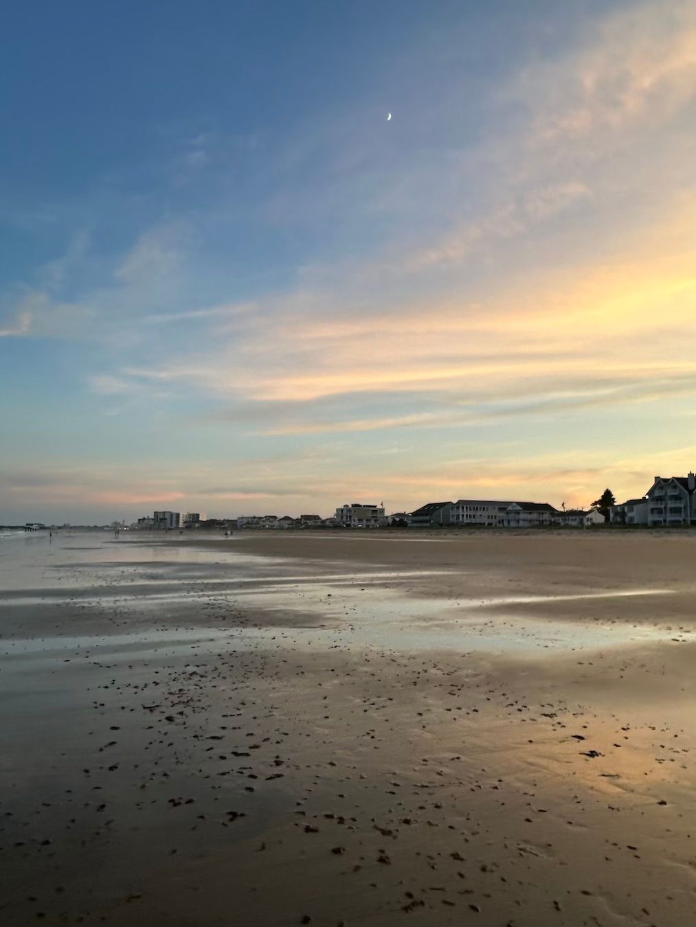 Wide sandy beach at sunset with coastal town skyline and crescent moon in colorful sky.