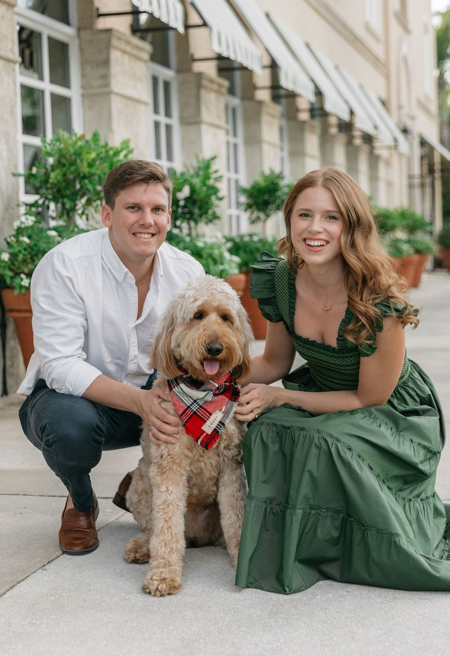 A couple sits on stone steps with their goldendoodle dog wearing a red bandana in an elegant European setting.