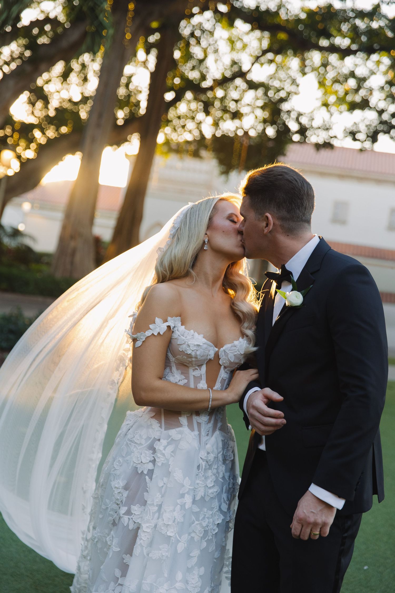 A romantic wedding moment captured at sunset with a flowing veil and elegant lace dress against a backlit garden setting.