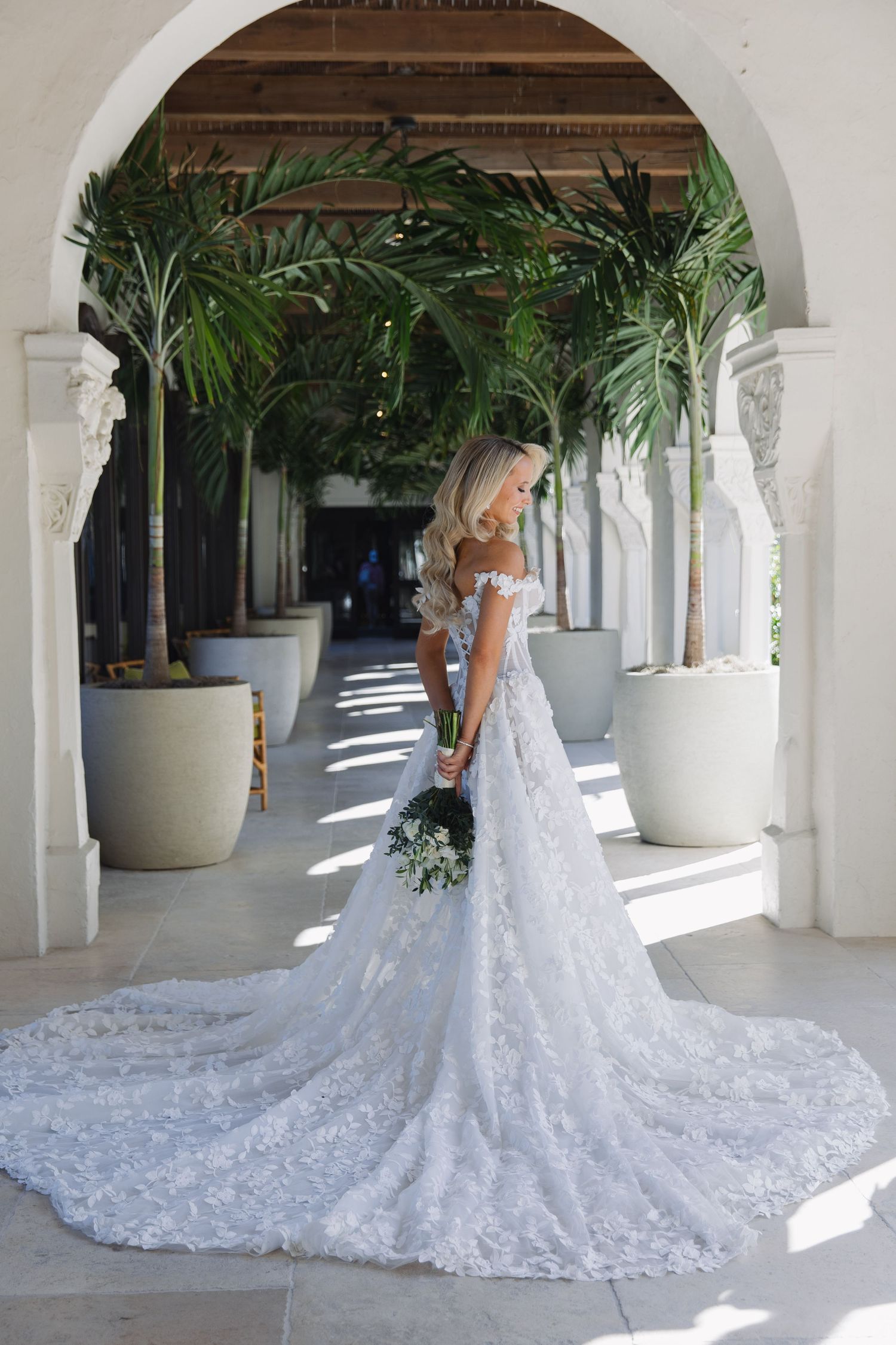 A stunning lace wedding gown with long train photographed in an elegant colonnade with tropical palm plants.