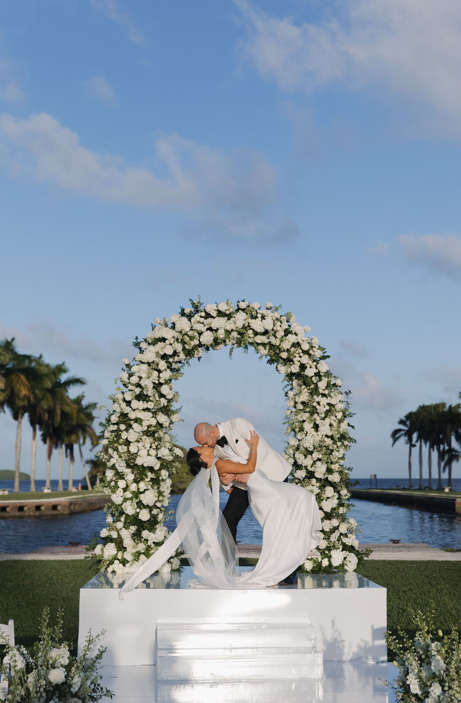 A romantic outdoor wedding ceremony under a white floral arch by the waterfront with palm trees in the background.