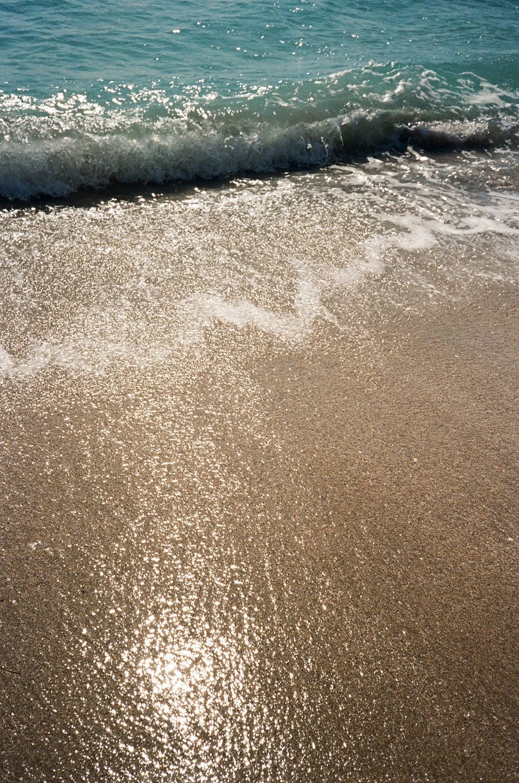 Sunlit waves crash onto a sandy beach creating sparkles in the shallow water.