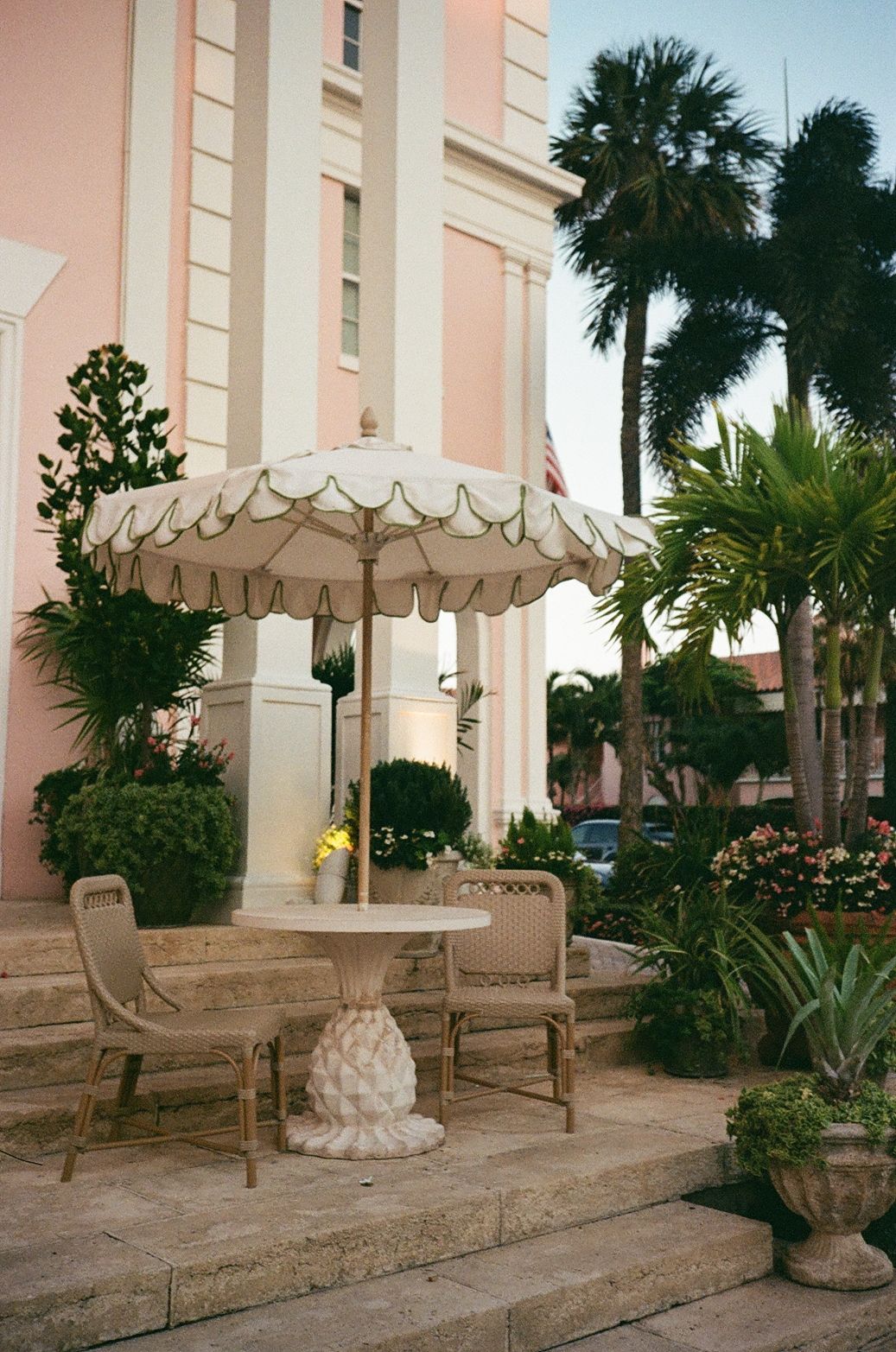 Outdoor seating area with elegant umbrella and benches against pink building with tropical landscaping.