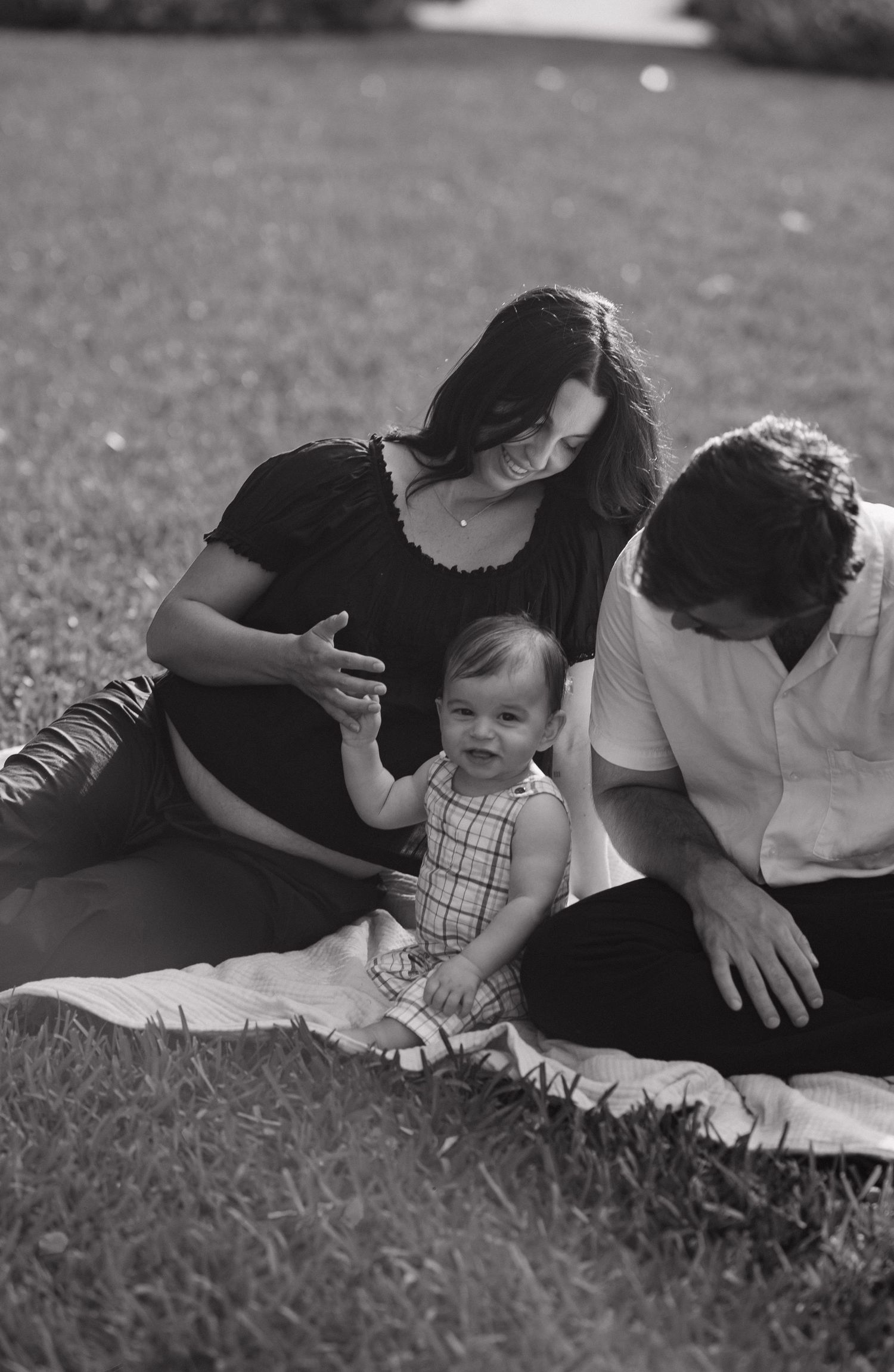 Black and white photo of a young family sitting on a blanket in the grass, with a baby between two parents.