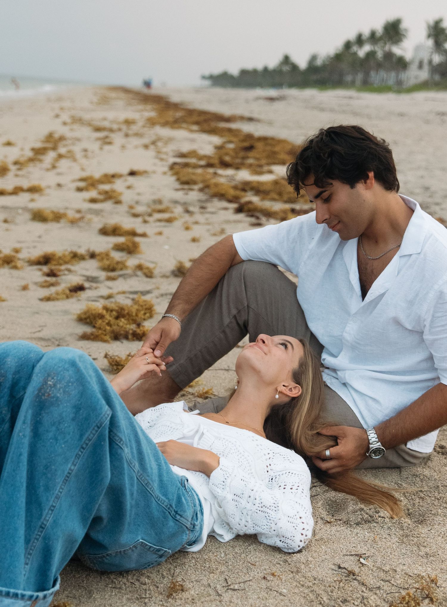 Couple relaxing together on sandy beach wearing casual white clothing.