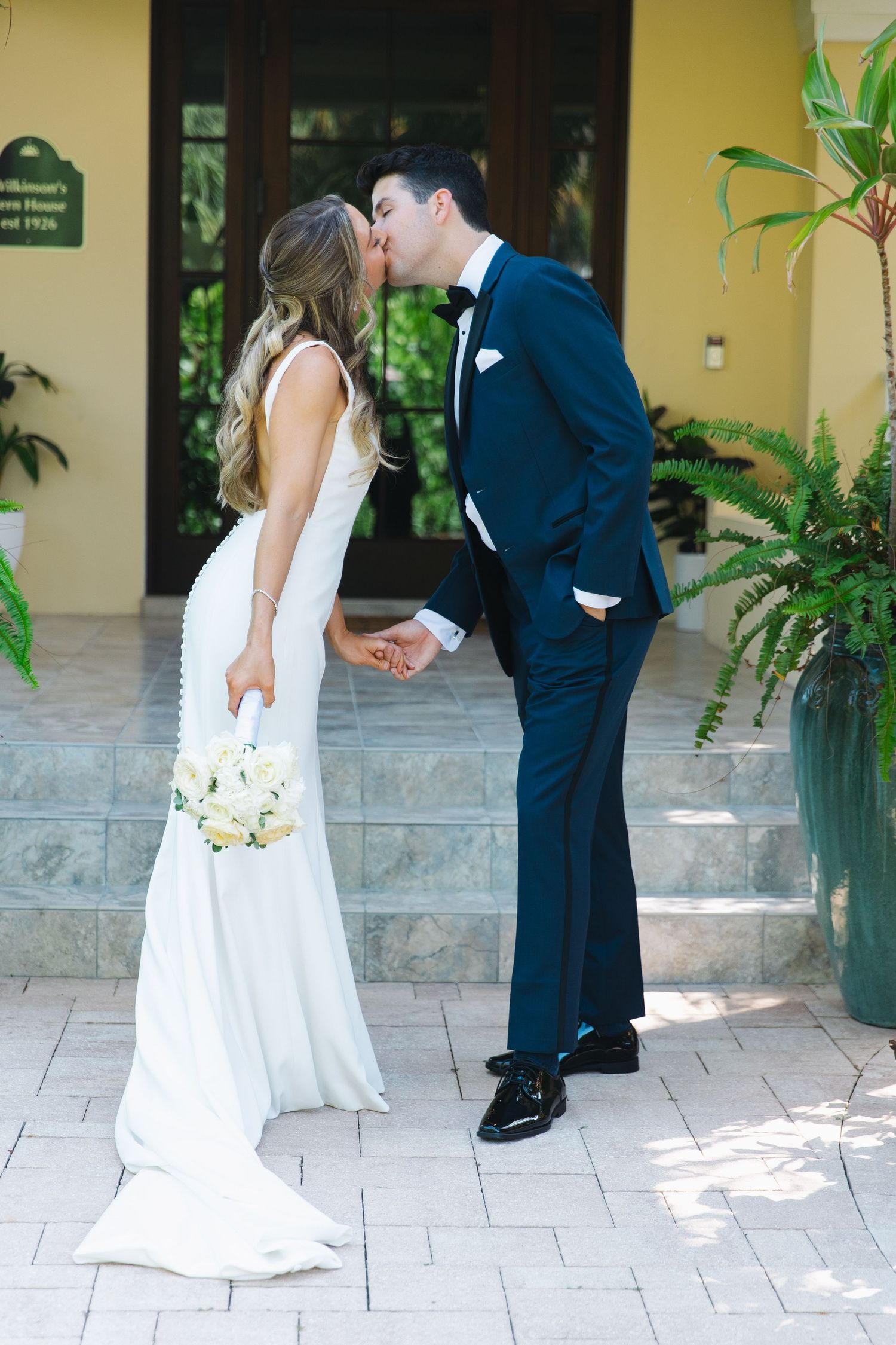 A couple in wedding attire share a tender moment while standing on a stone patio surrounded by greenery and planters.