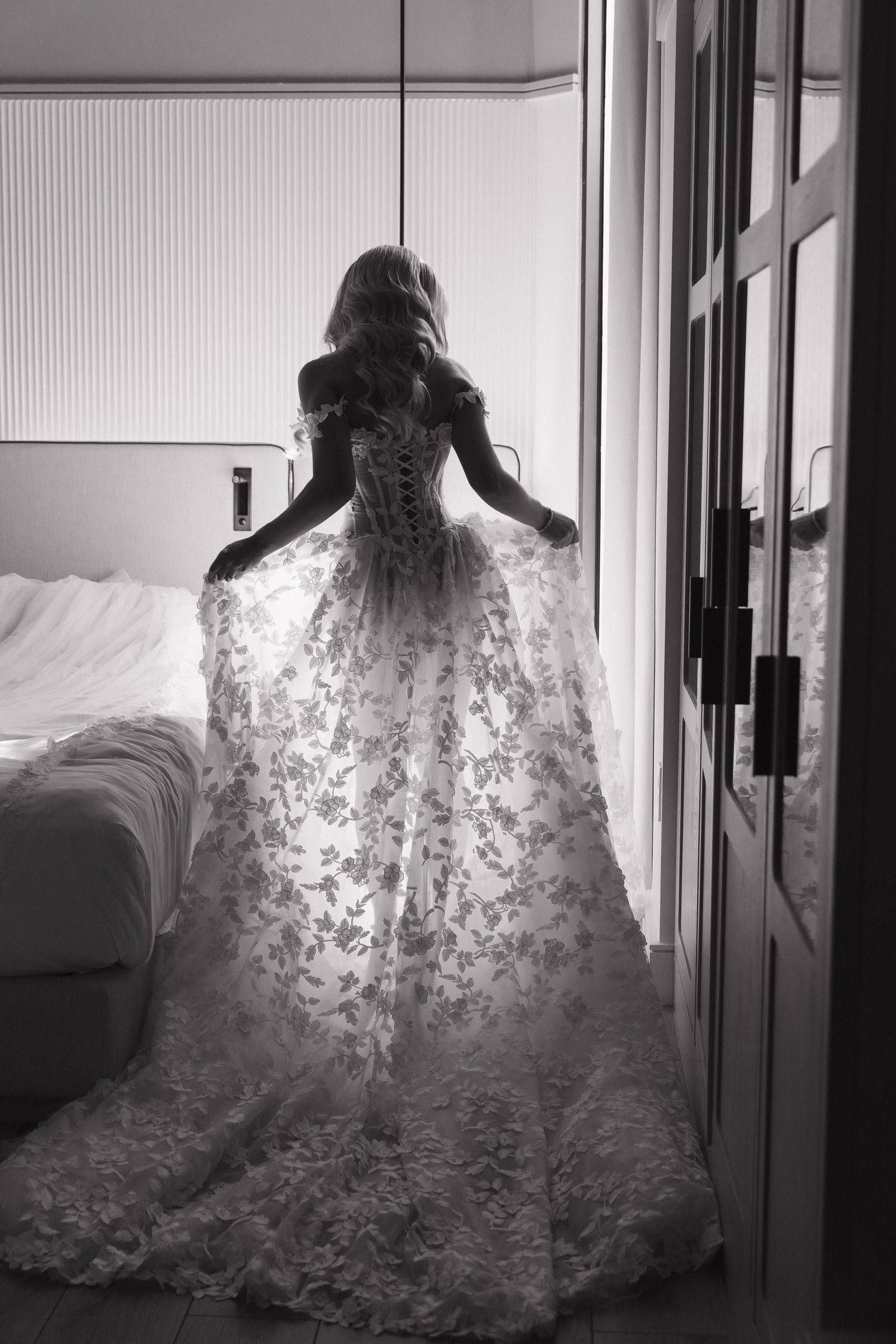 Silhouette of a bridal gown with delicate floral lace patterns illuminated from behind in a corridor.