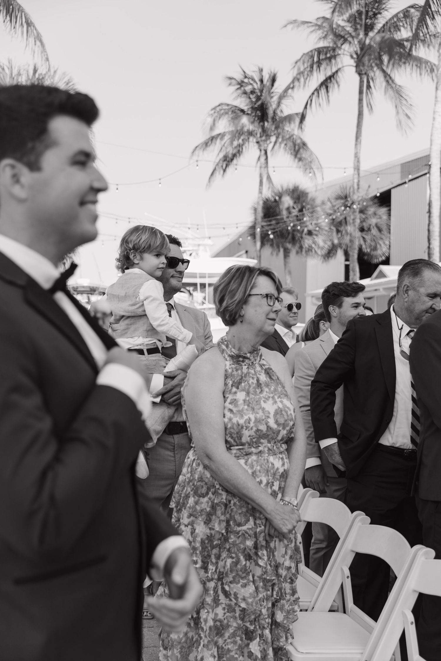 Black and white photo of guests at an outdoor event with palm trees, featuring people in formal attire gathered together.