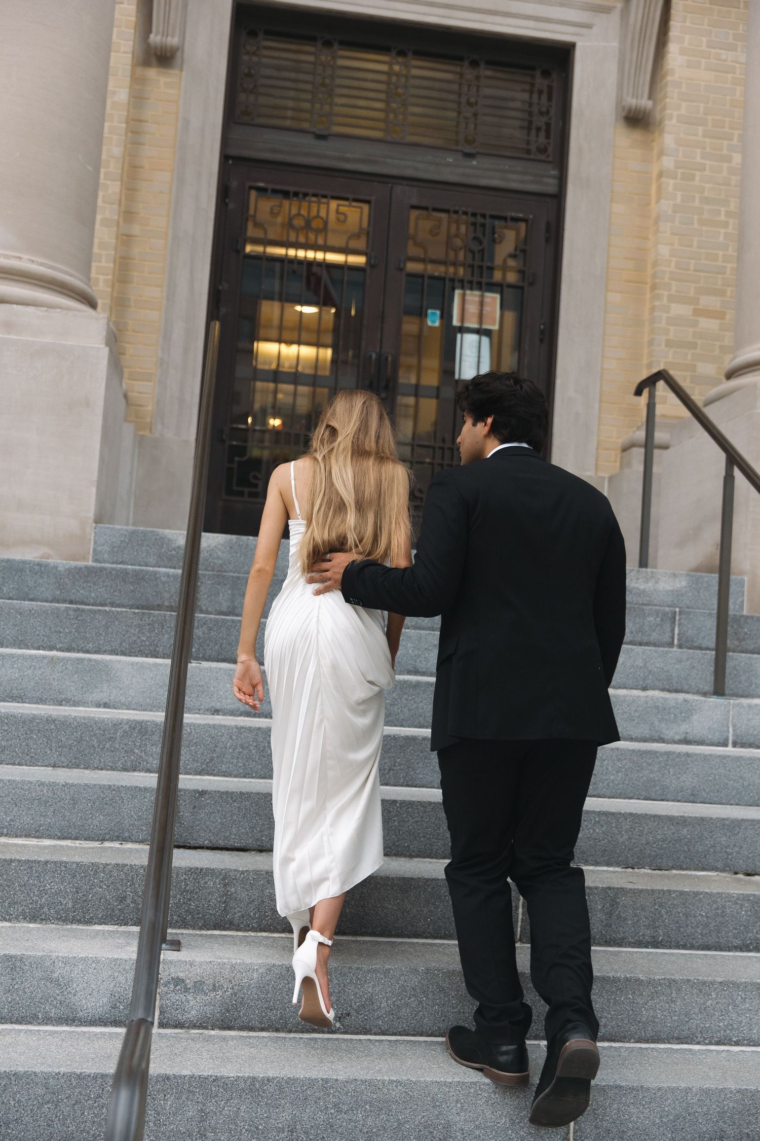 Couple ascending stone steps together, with the bride in white and groom in black at a classic building entrance.