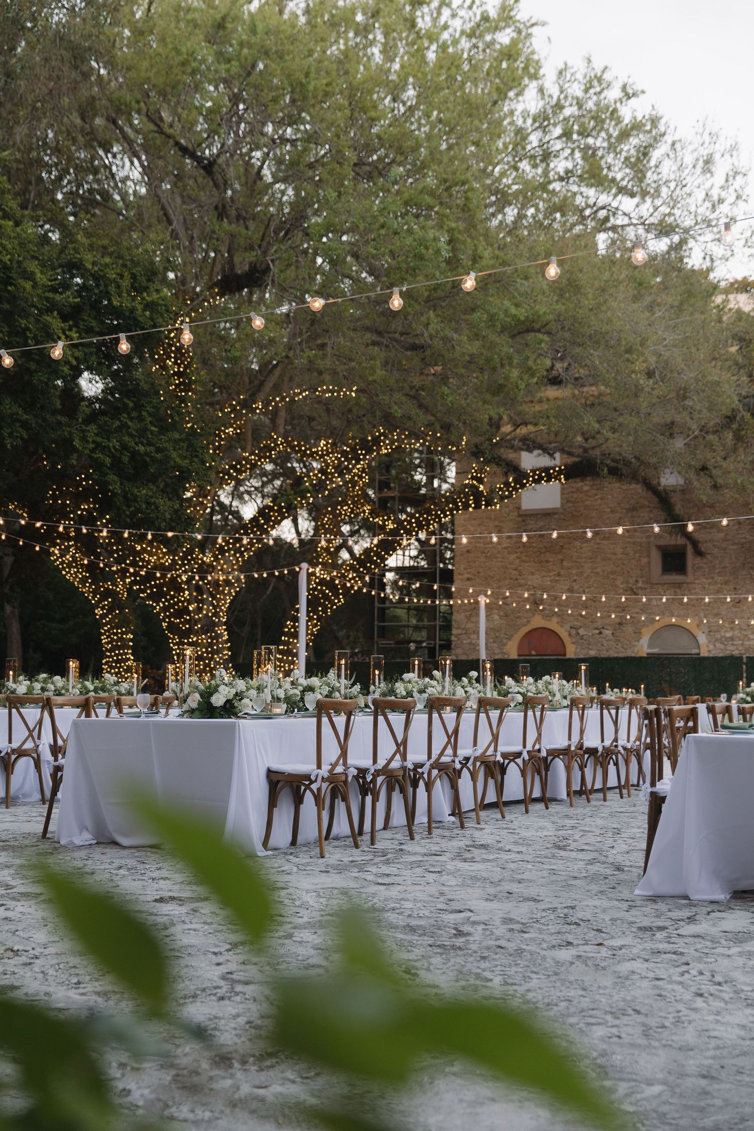 Outdoor wedding reception setup with string lights, white tablecloths and wooden cross-back chairs under illuminated trees.