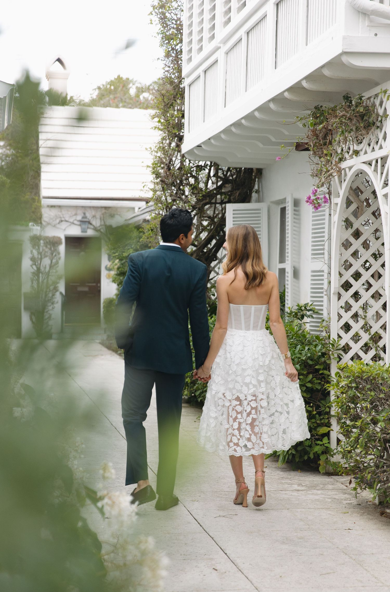 A romantic couple walking along a white garden path with lattice architecture and blooming flowers.