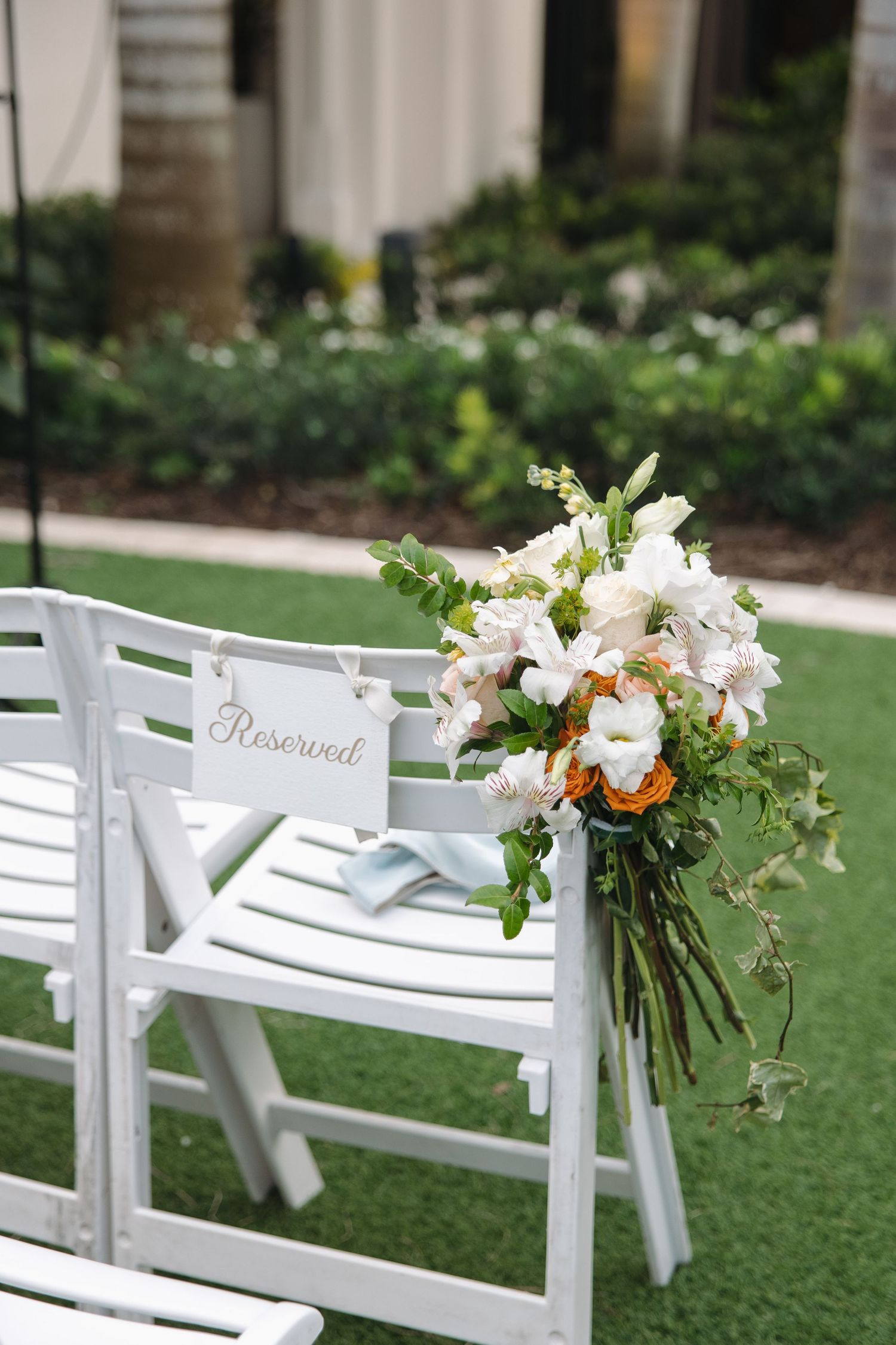 White folding chairs with 'Reserved' sign and floral arrangement set up for outdoor garden wedding ceremony.