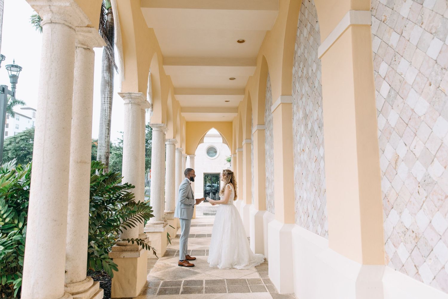 Wedding couple stands in an elegant covered walkway with cream colored columns and textured walls on a bright sunny day.