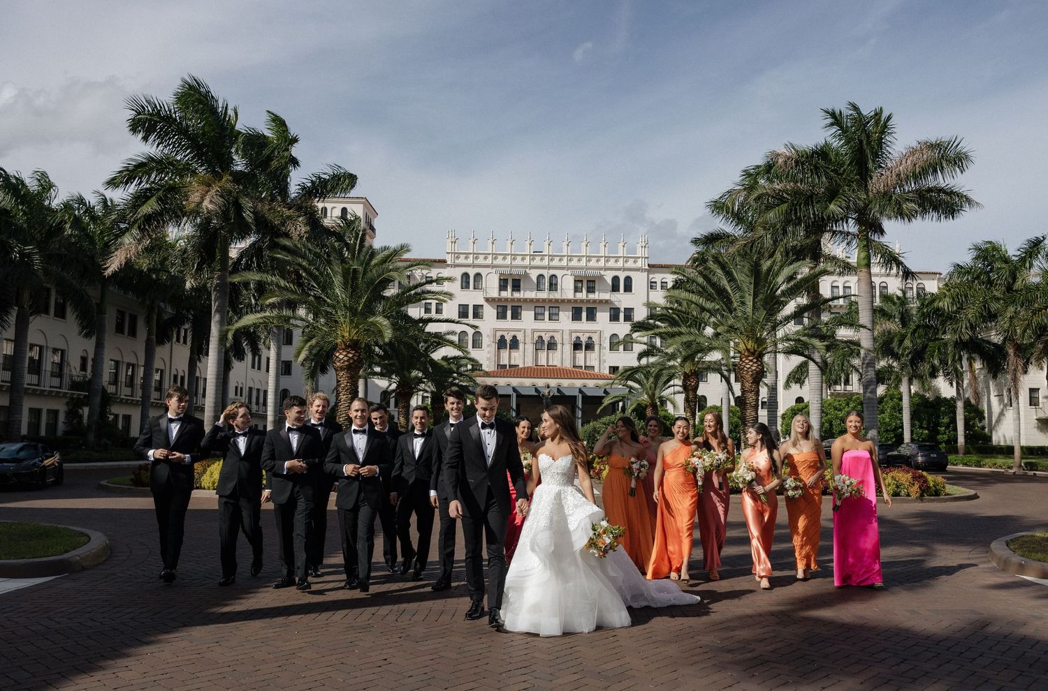 Wedding party members line up for formal photos in front of palm trees at a tropical venue.