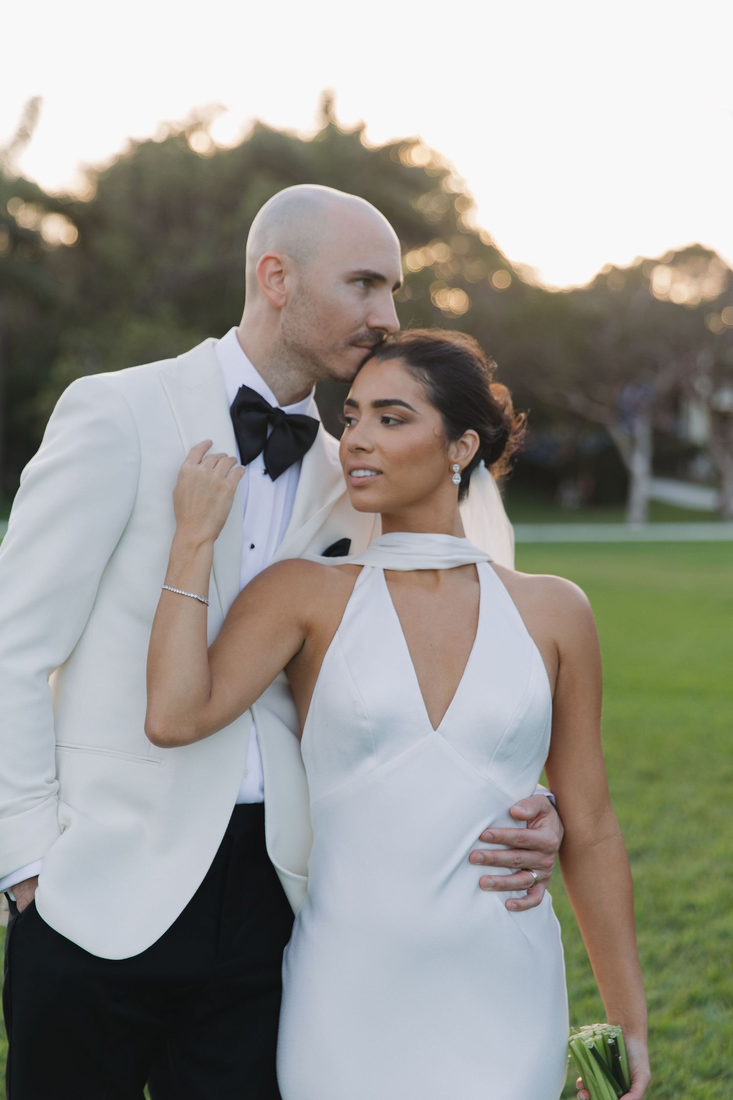 Romantic wedding couple in elegant white dress and black tuxedo embrace on a grassy lawn at sunset.