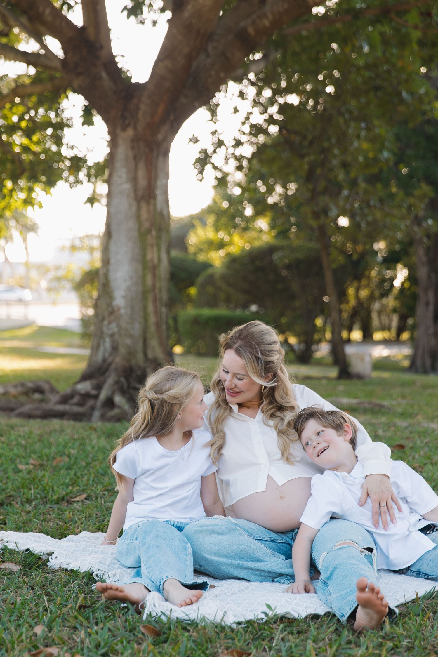 Pregnant woman sitting on blanket in park with two children in white tops and jeans, smiling under large tree.