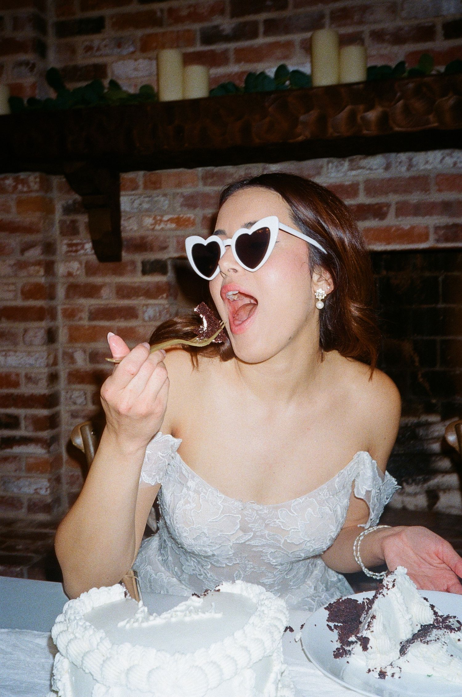 A person in heart-shaped sunglasses and white dress enthusiastically eats wedding cake at a rustic brick venue.