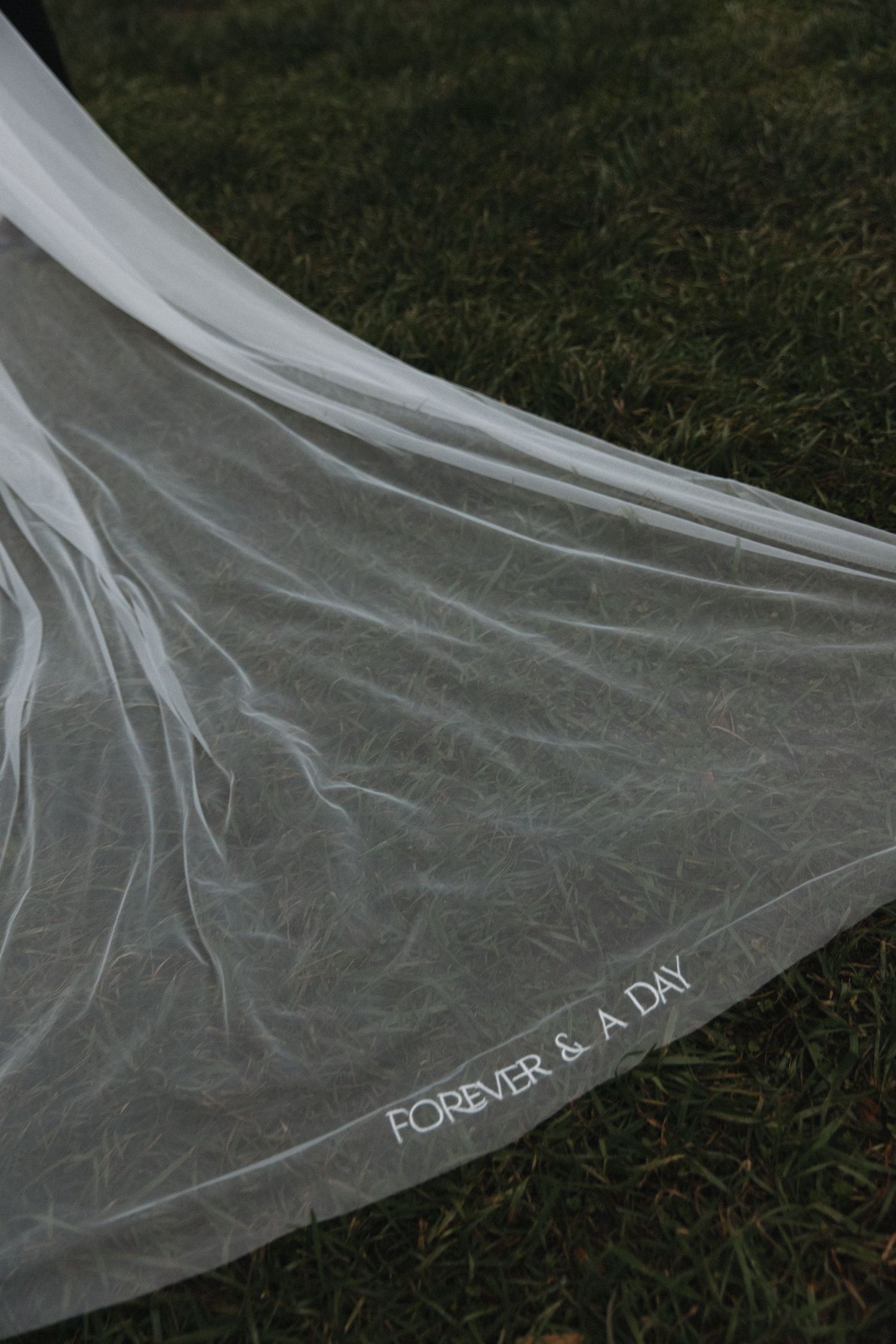 A delicate wedding veil spread on grass with 'FOREVER & A DAY' text embroidered along the edge.