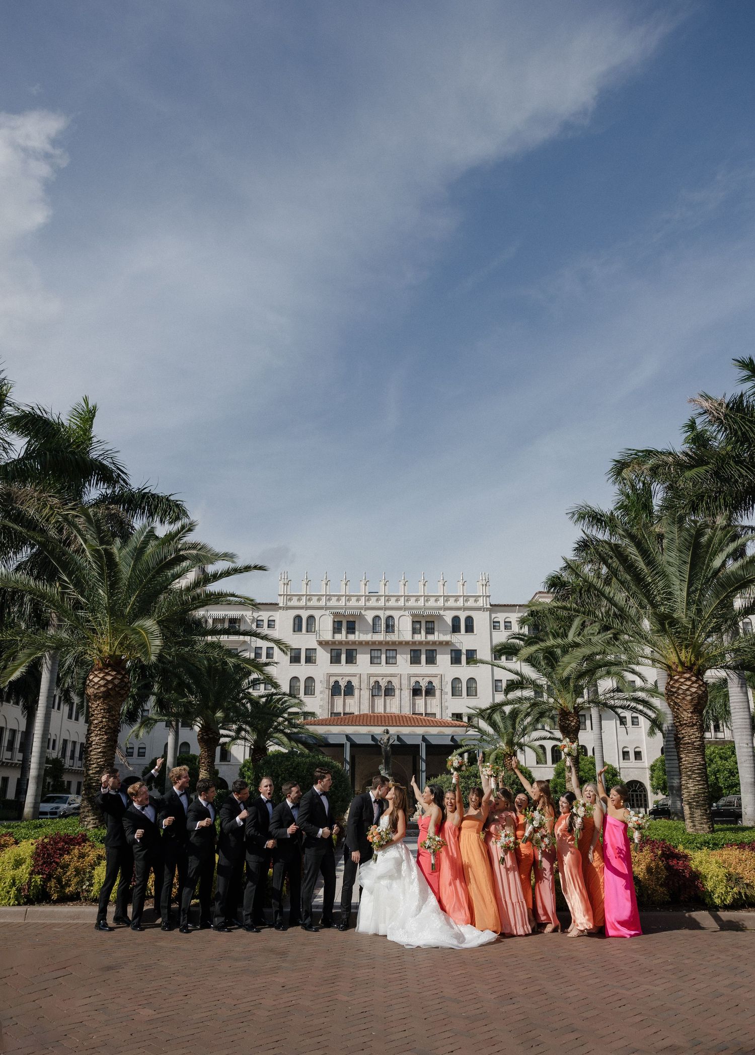 Wedding group photo with groomsmen in black and bridesmaids in pink dresses at tropical resort.