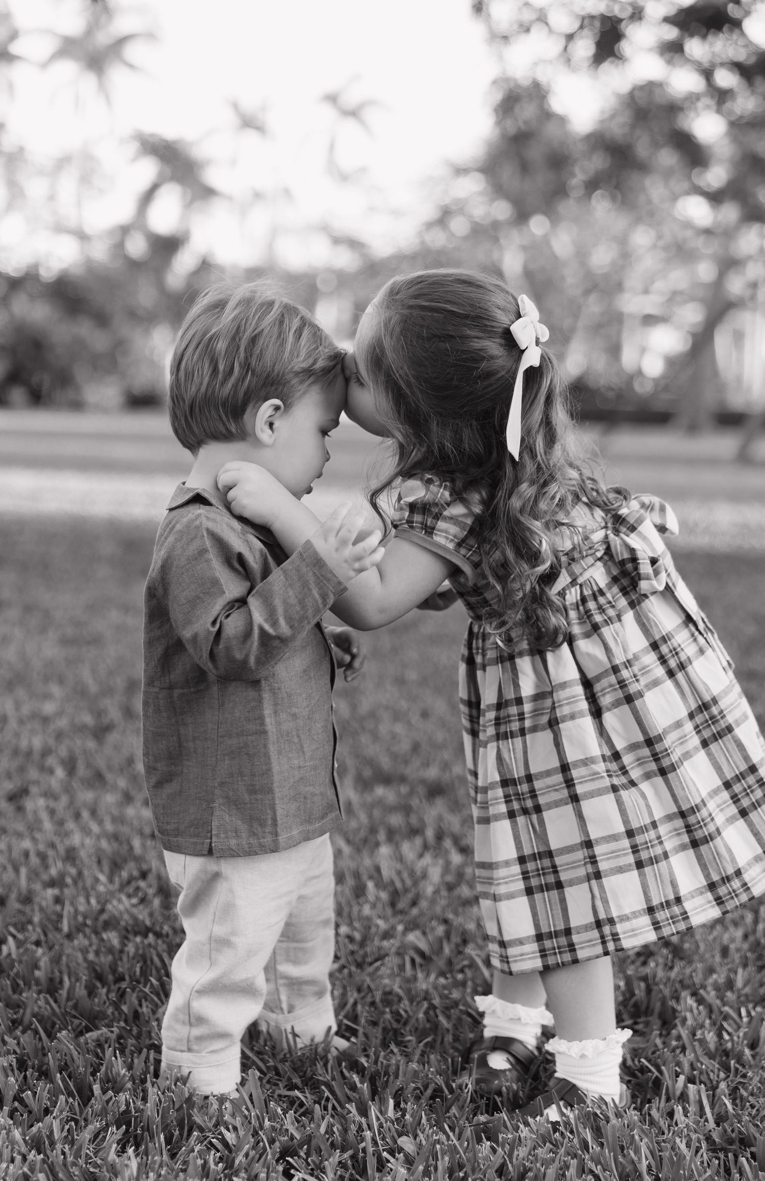 Two children sharing a sweet kiss while standing in grass wearing plaid dress and casual clothes.