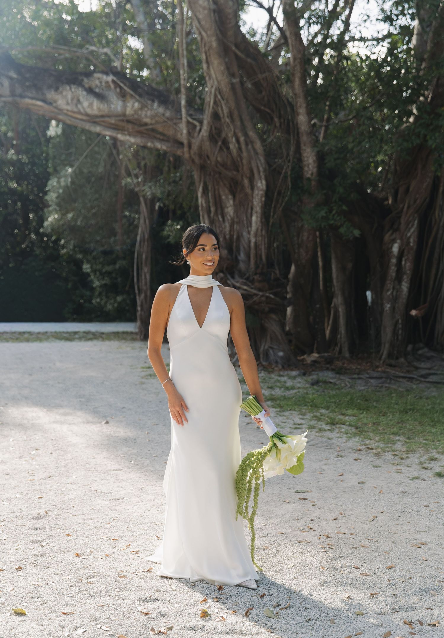 A person in a white halter dress poses against a backdrop of lush trees and sandy ground at an outdoor location.