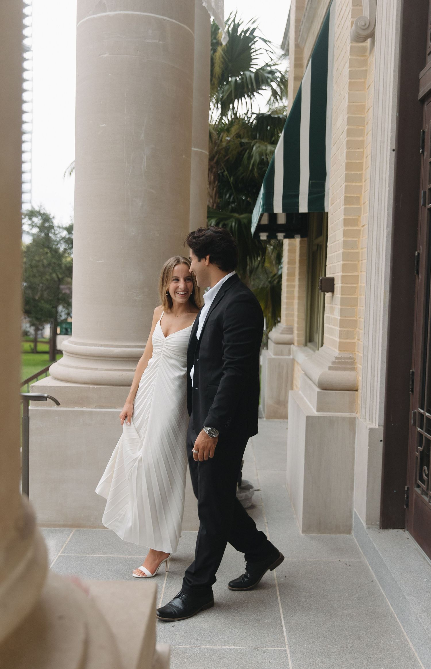 Couple in formal wear playfully interacting along a columned walkway.