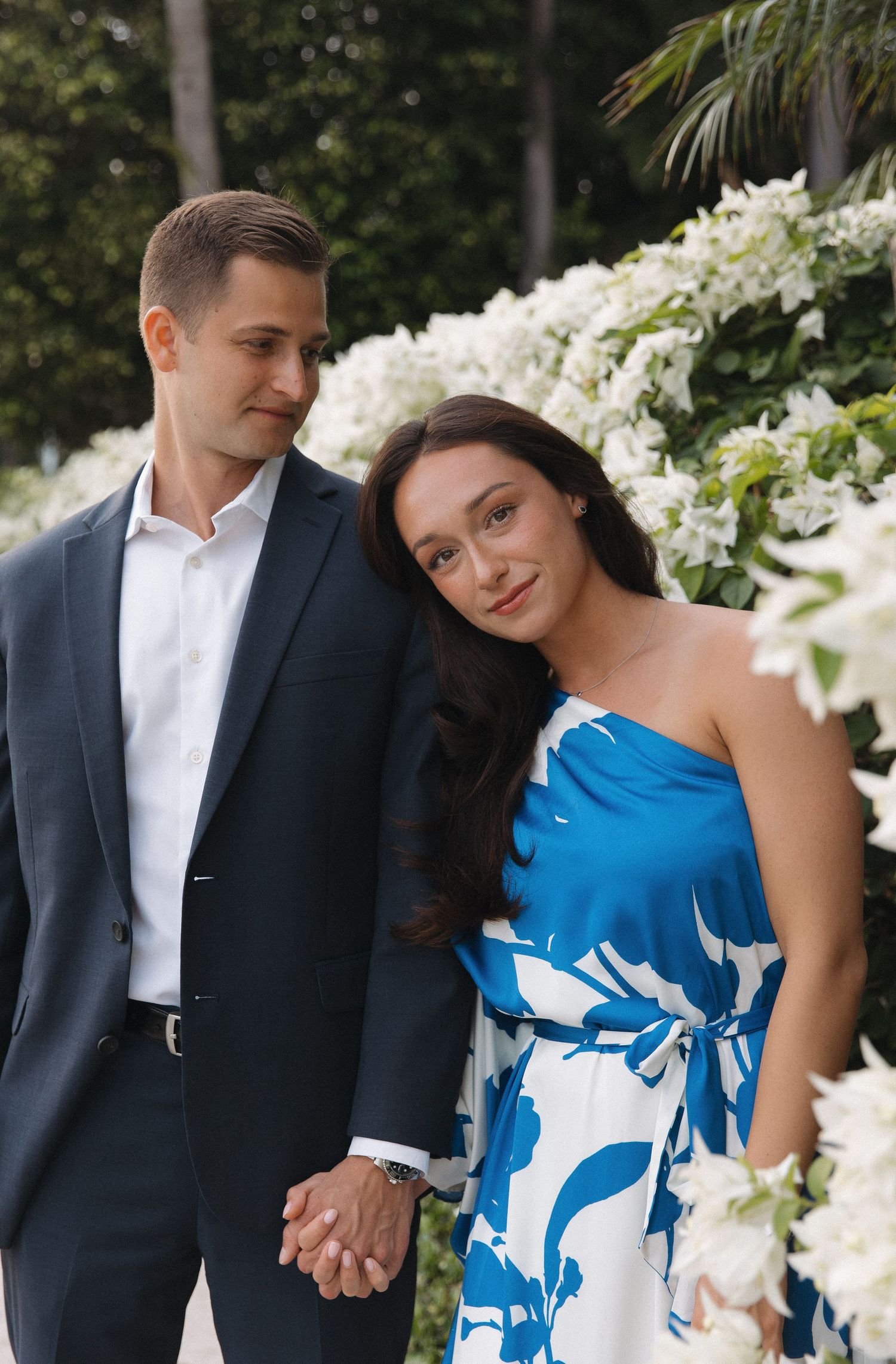 A couple in formal attire poses together against a backdrop of white flowers in a garden setting.