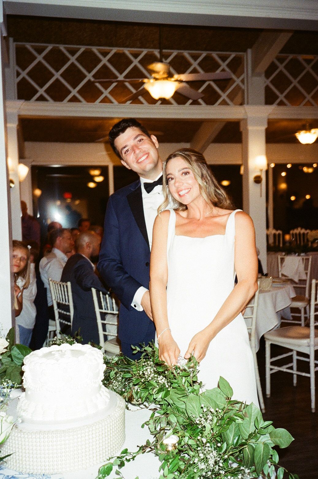 A couple celebrating at their wedding reception stand beside an elegant white wedding cake decorated with greenery and flowers.