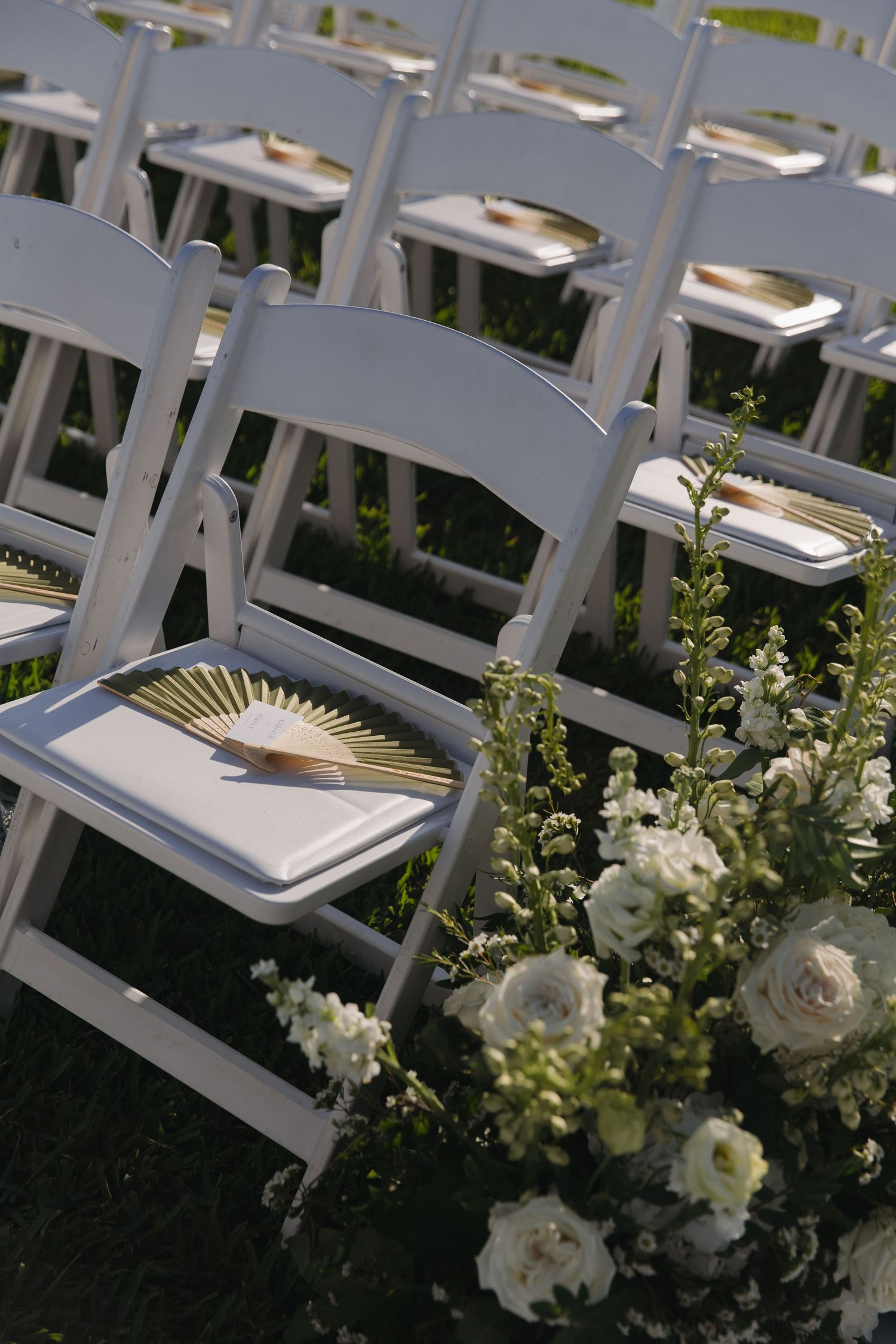 White folding chairs decorated with delicate white floral arrangements line up for an elegant outdoor ceremony.