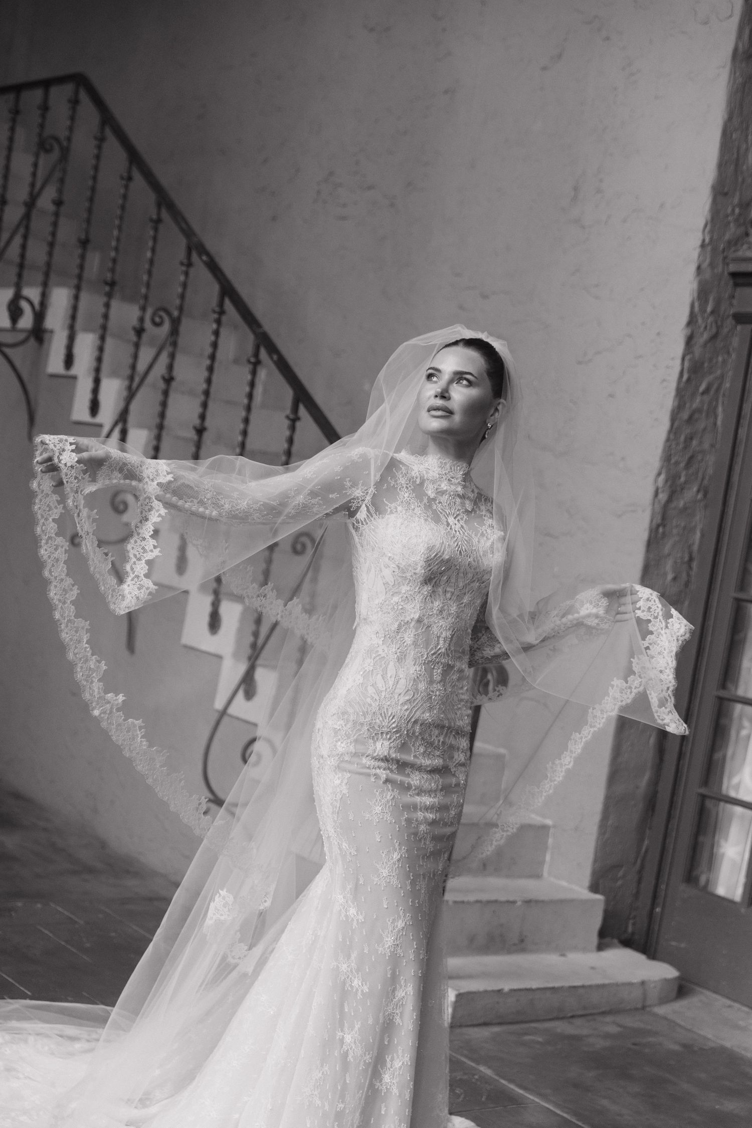 A bride in an elegant lace wedding gown with flowing veil poses gracefully on a staircase in black and white photography.