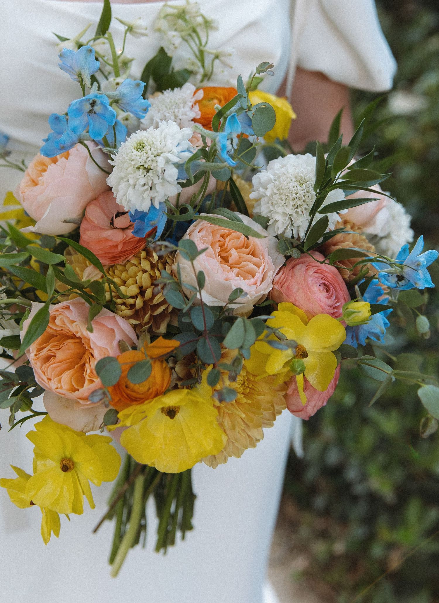 Delicate wedding bouquet featuring peach roses, yellow daffodils, and blue delphinium flowers against a white background.